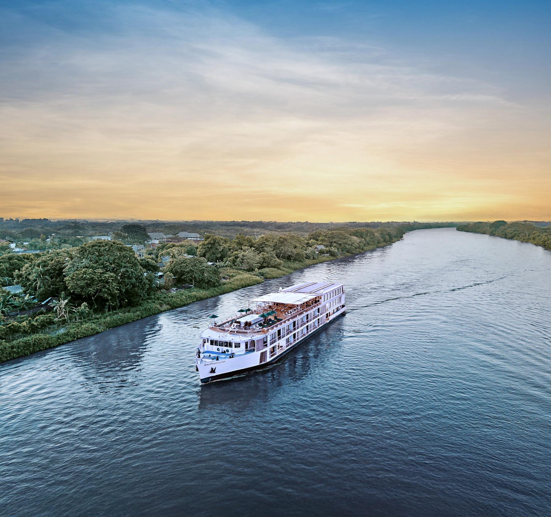 AmaMagdalena from the air at golden hour — the 60-guest ship mid-river on the Magdalena, wake trailing behind, forest on both banks.