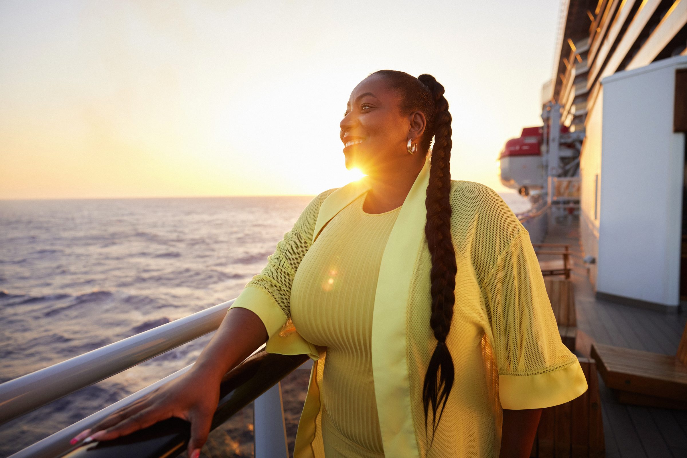 A woman in a pale yellow knit jacket leans on a ship's rail at sunset, smiling out over the open sea, the promenade deck and a lifeboat stretching away behind her.