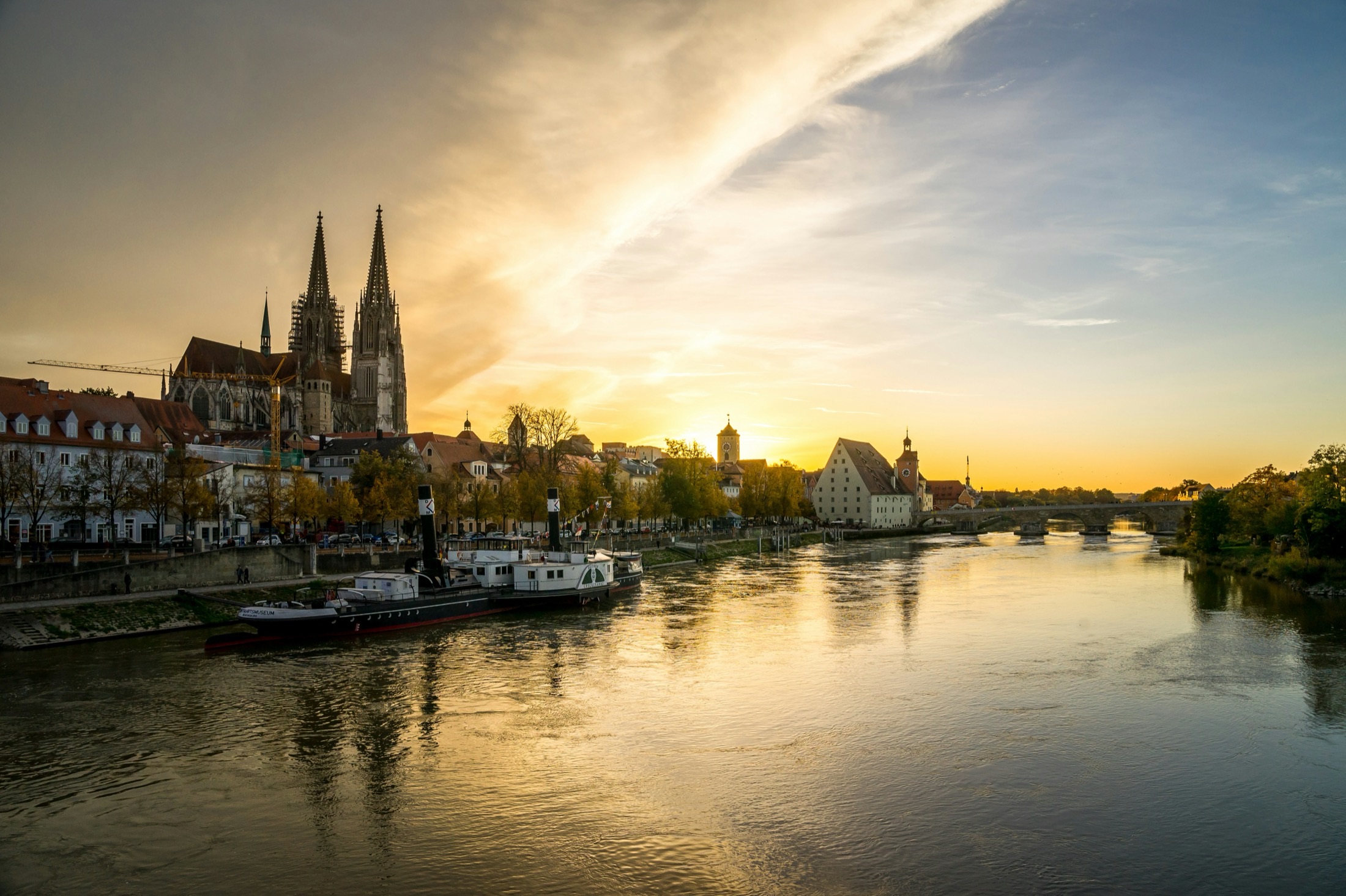 The Danube at Regensburg, Germany, at golden hour — cathedral spires, the medieval Stone Bridge, and a moored river vessel.