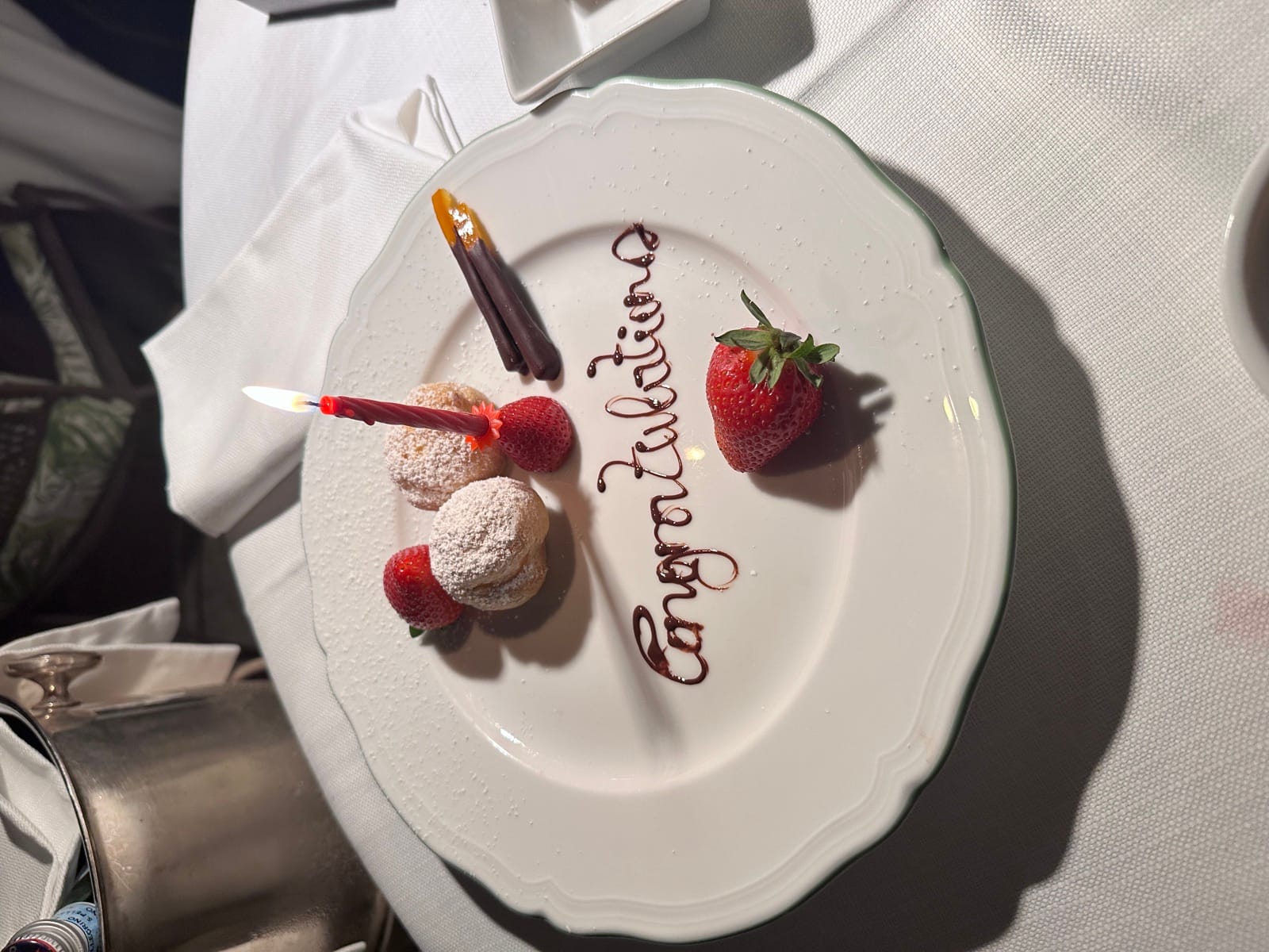 A dessert plate at Le Jardin in the Hotel de Russie garden with 'Congratulations' written in chocolate, strawberries, powdered-sugar bignè, and a lit candle.