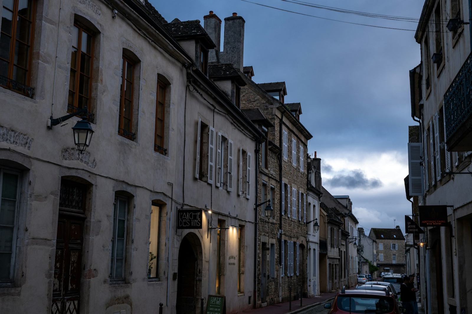 A quiet cobblestone street at dusk in Beaune, Burgundy — warm light glowing in the windows of old stone buildings.
