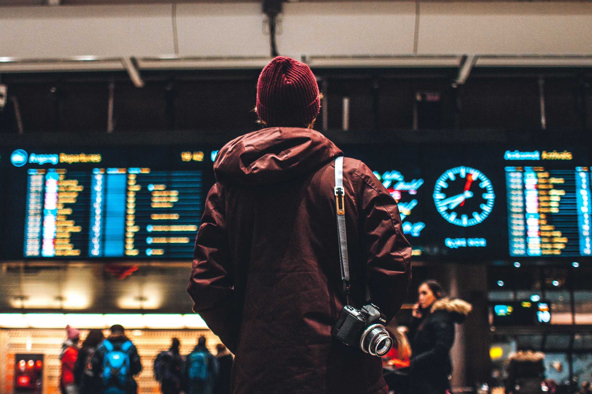 A traveler in a dark coat and wool hat stands with his back to the camera beneath the glowing departures and arrivals boards of a busy airport, a clock reading past the hour above him.