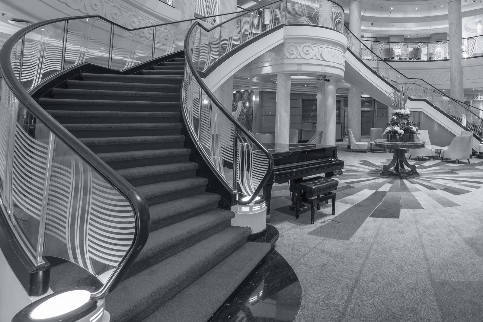 The Grand Lobby of Queen Mary 2 in black and white: a sweeping staircase with etched-glass balustrades, a grand piano at its foot, the lobby floor radiating out beyond.