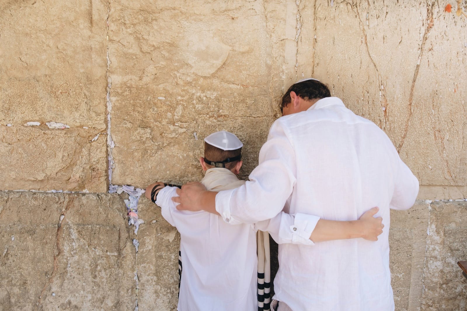 A father in white linen holds his son close at the Western Wall in Jerusalem; both wear kippot, the son in tefillin, prayer notes tucked into the limestone seams.