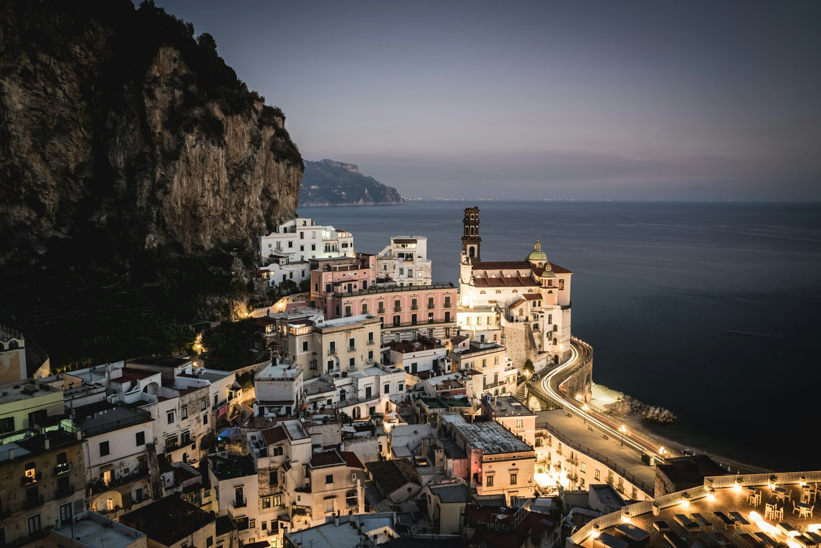Atrani at dusk, lights coming on in stacked pastel houses folded into the cliff above a small beach.