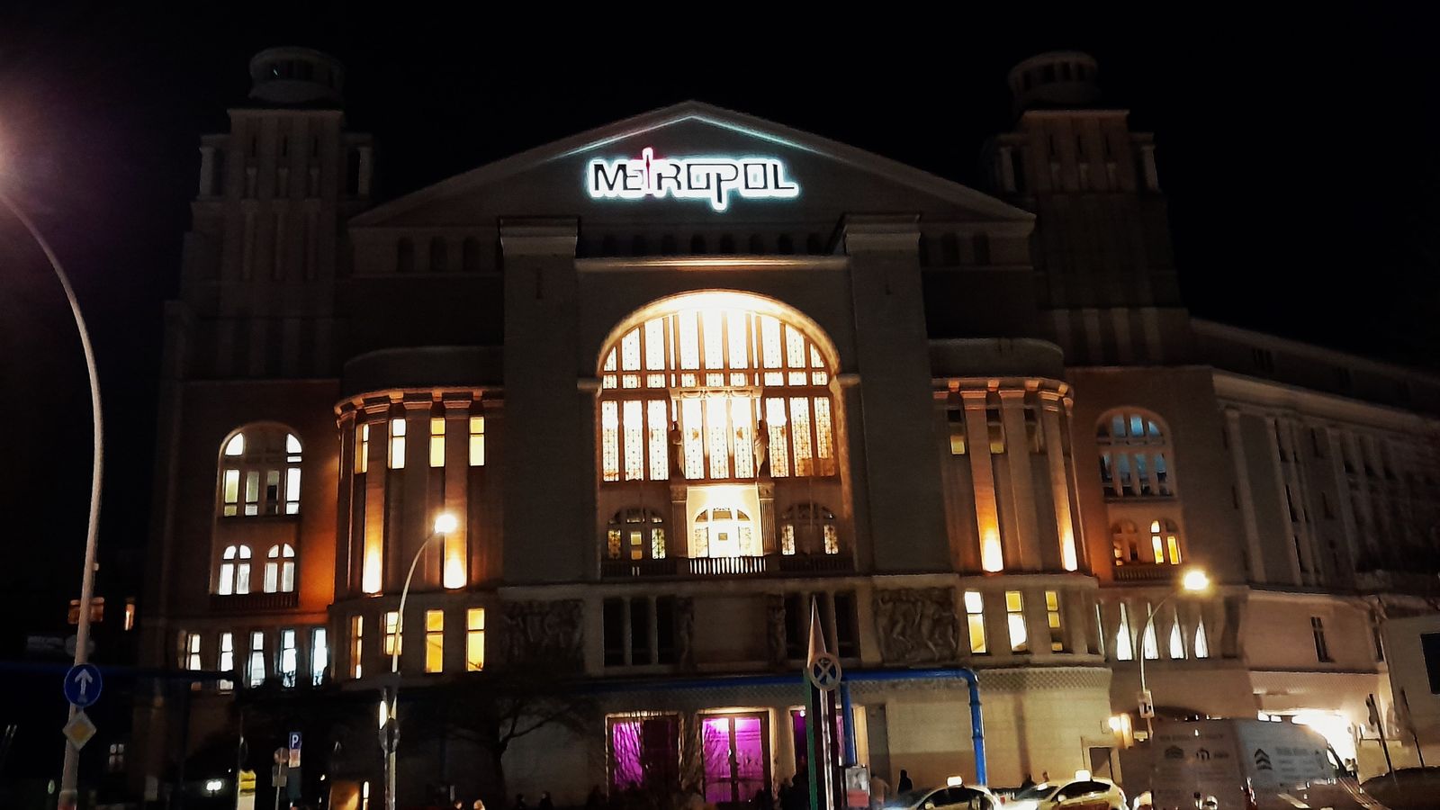 The Metropol at Nollendorfplatz in Berlin lit at night — neon sign above the facade, the great arched window glowing amber.