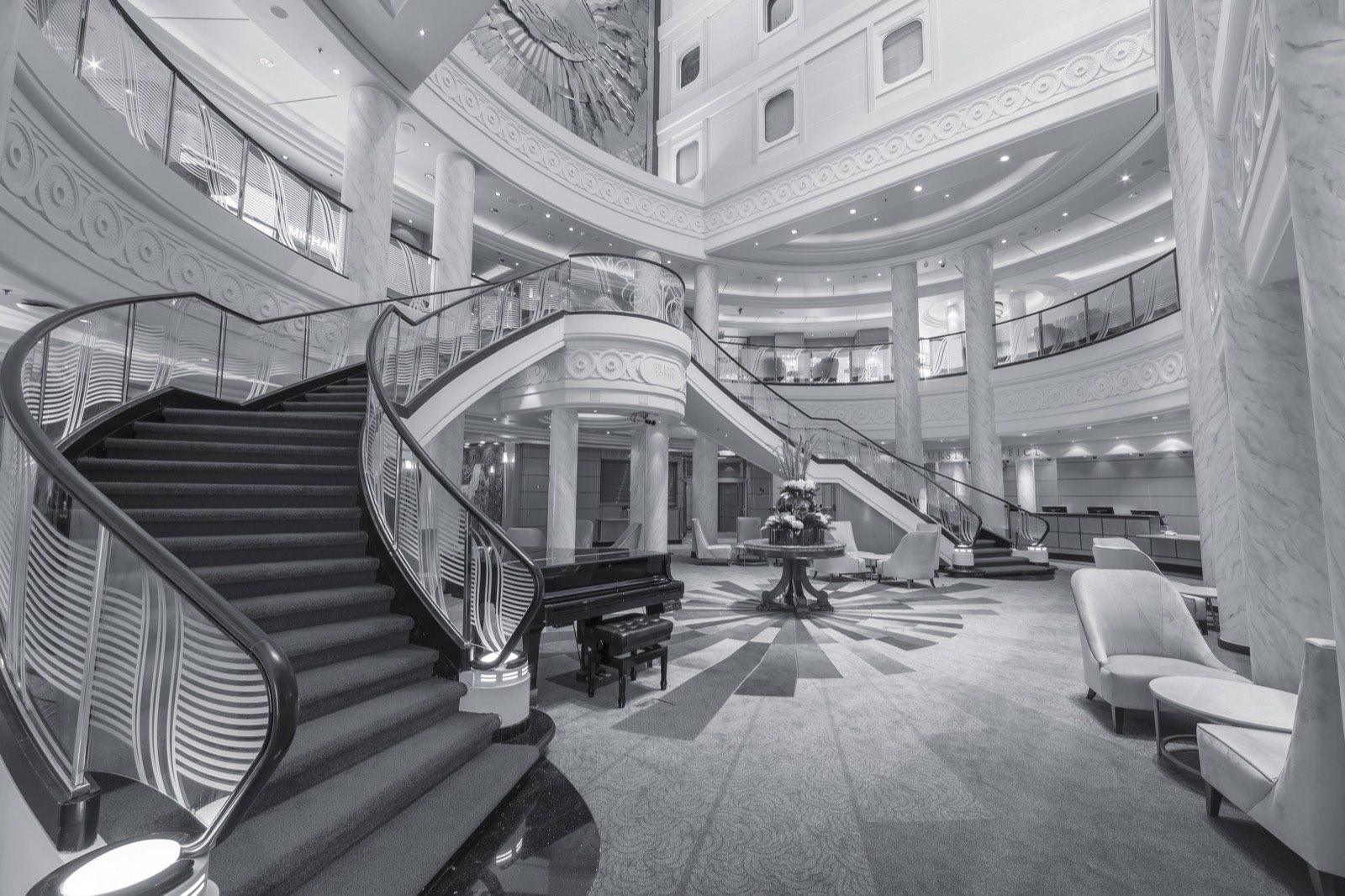 The Grand Lobby of Queen Mary 2 in black and white — a double staircase curving down to a marble floor, a grand piano at its foot, under a domed ceiling.