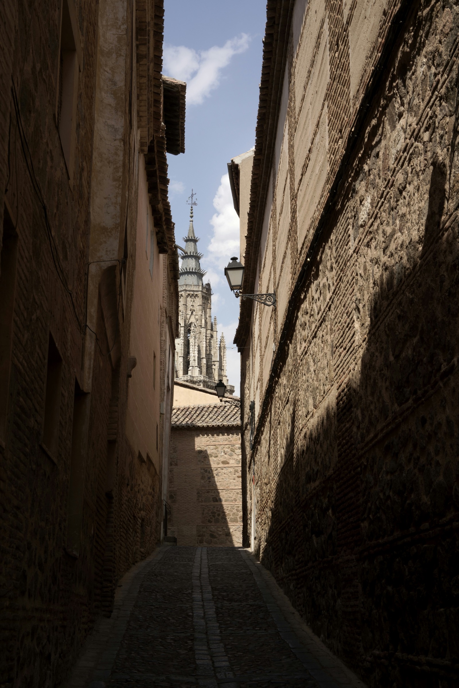 A narrow, empty stone lane climbs through Toledo's old town toward a distant bell tower — the kind of quiet alley that threads the city's medieval Judería.