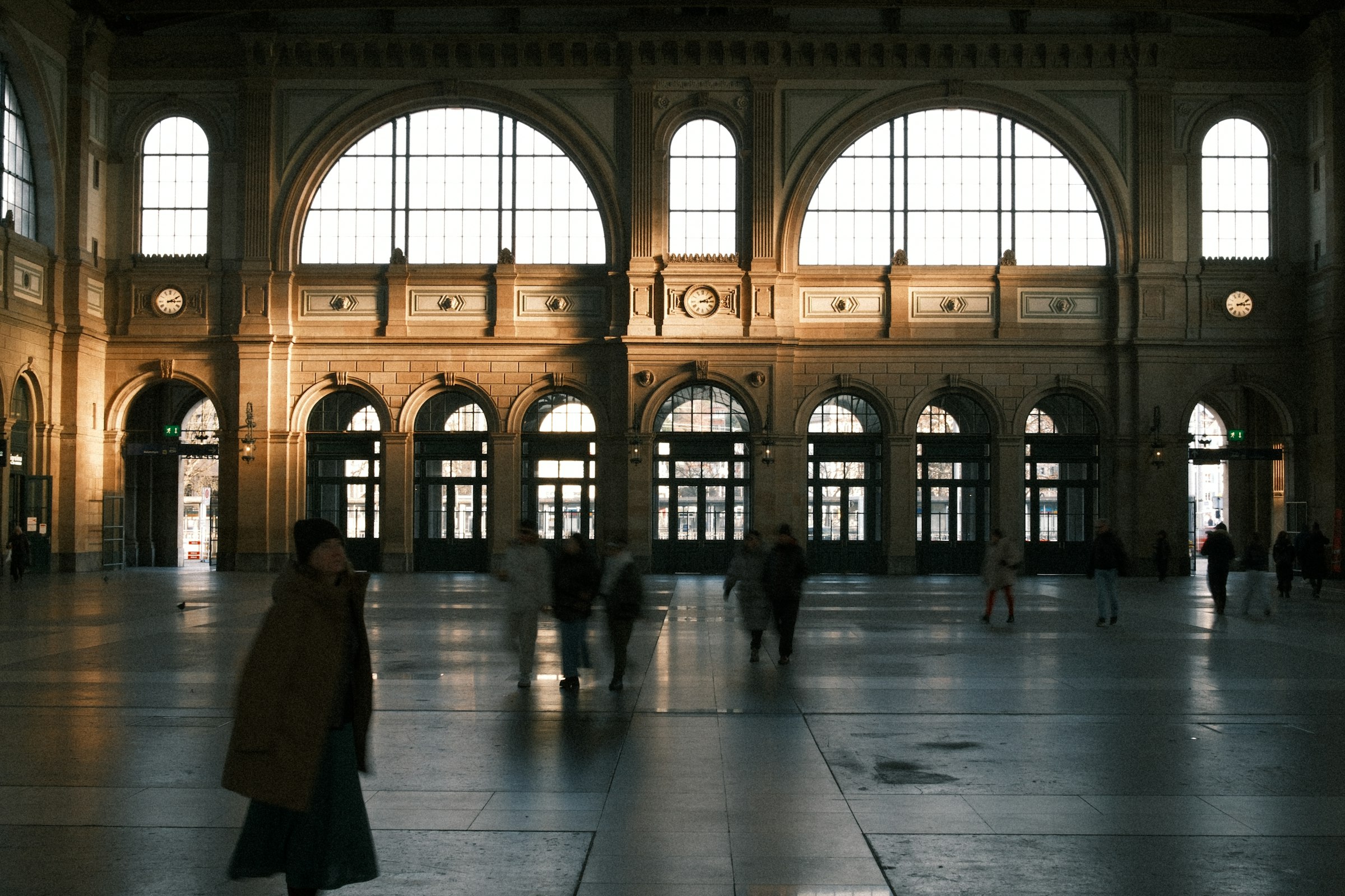 A grand, sunlit train station hall with travelers passing through — the great terminus where timing is everything.