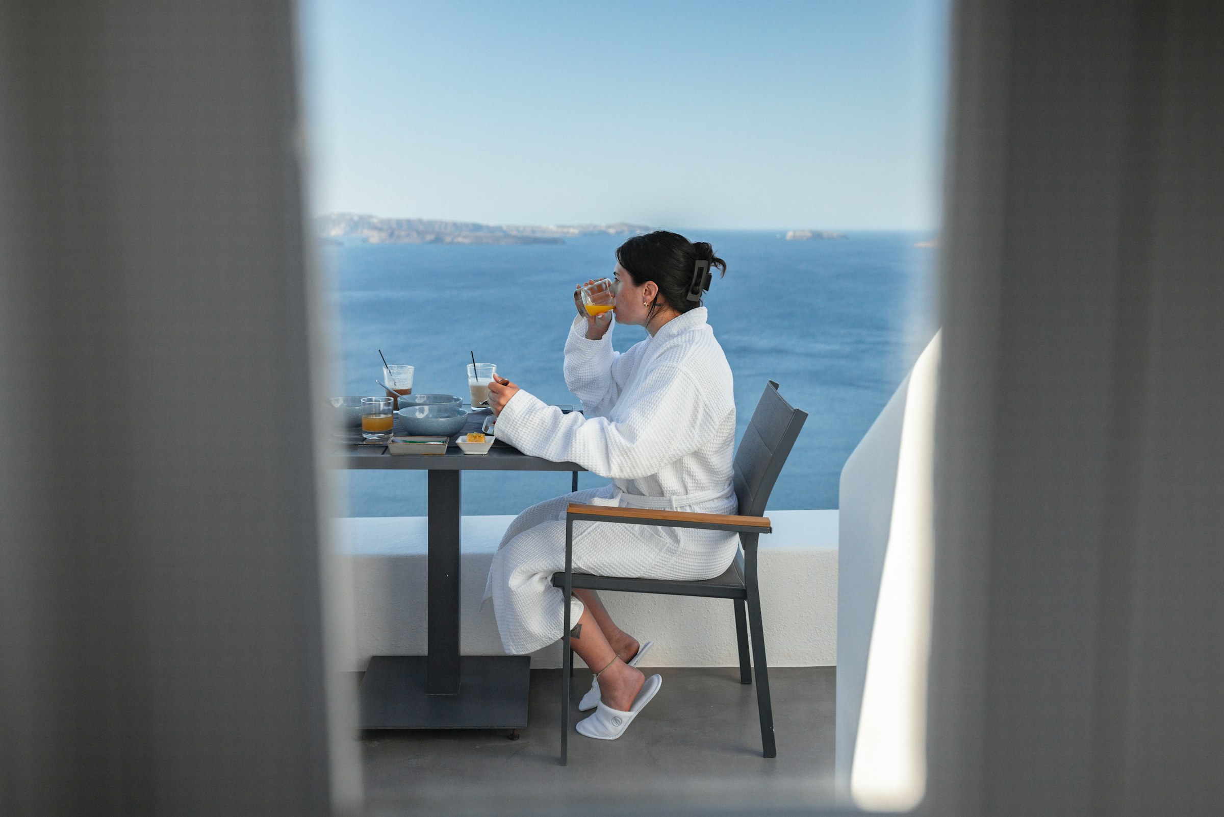 A woman in a white hotel bathrobe takes breakfast on a sunlit balcony above a deep blue sea, a mountainous coastline hazy in the distance.