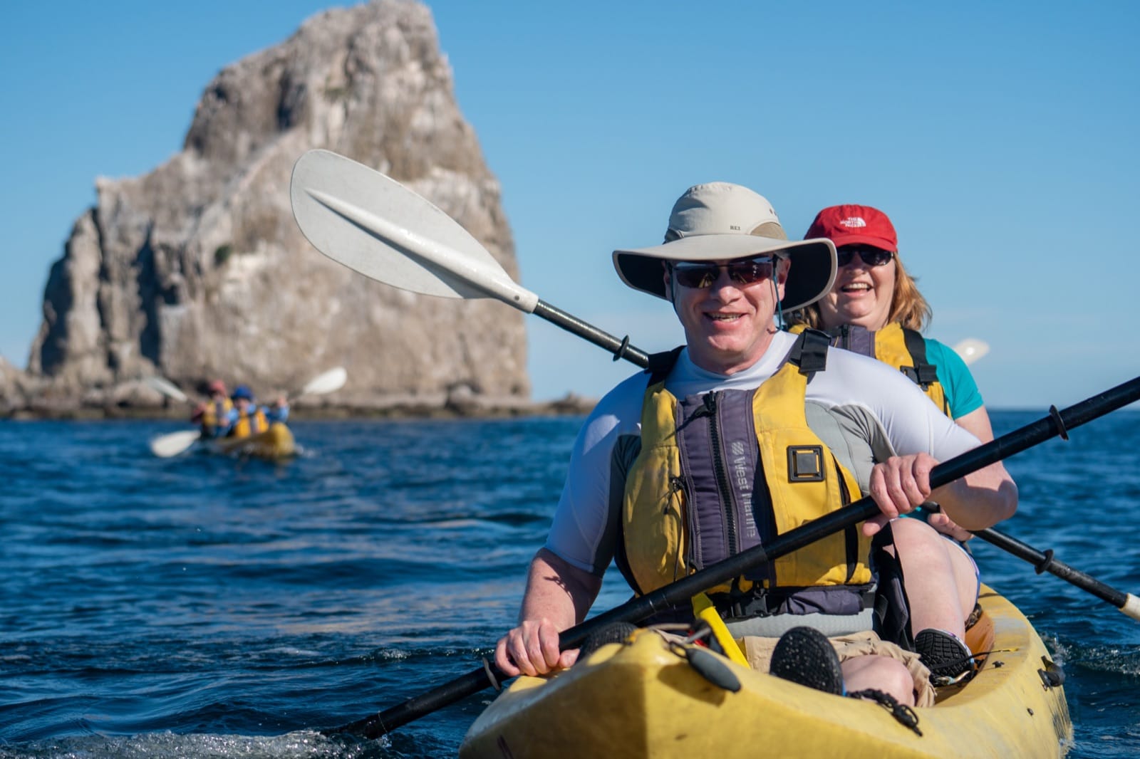 Two guests in a yellow kayak in Bahía Agua Verde, with a dramatic rock formation rising from the water behind them.