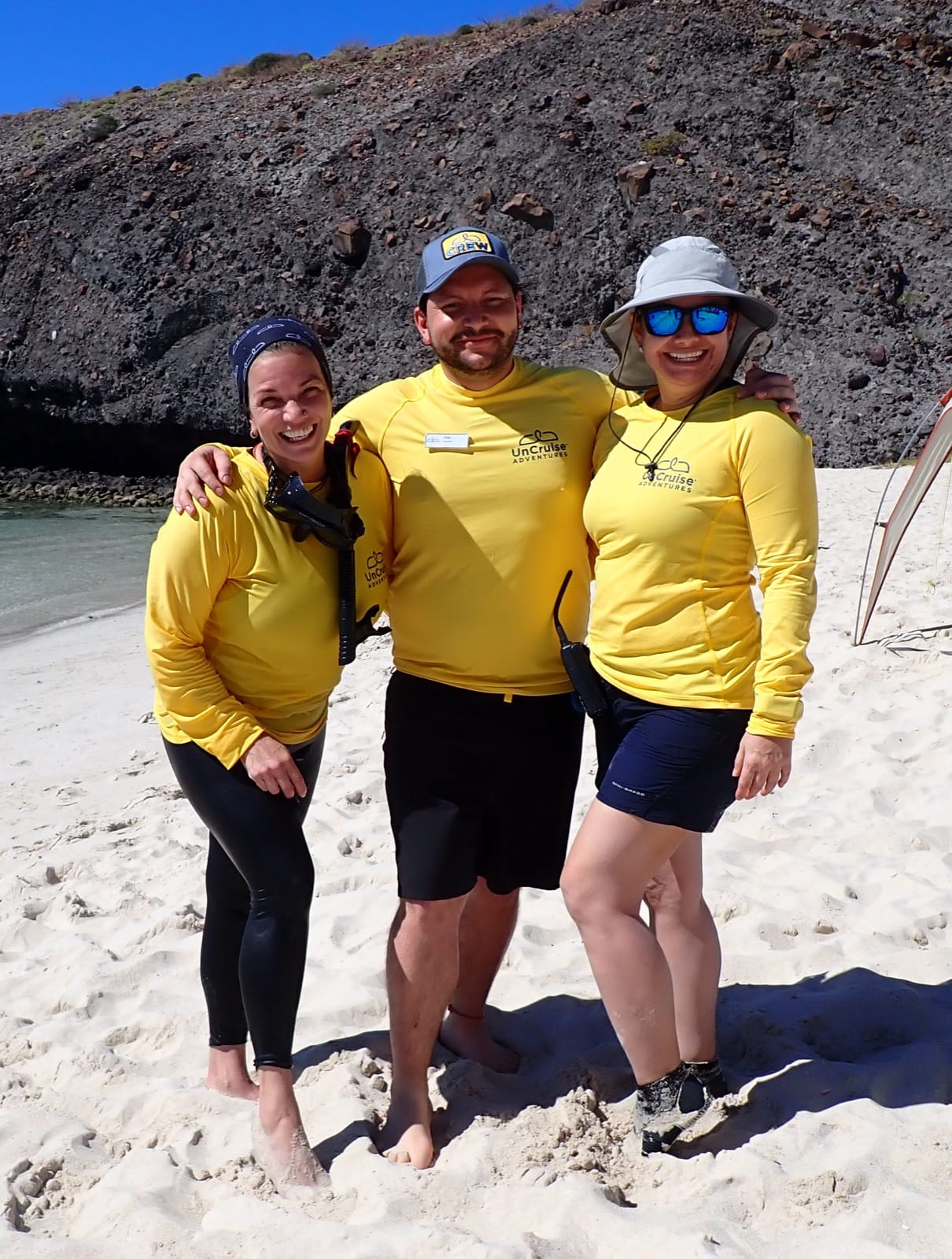 Three Safari Voyager crew members in yellow shirts standing together on a Baja beach, smiling at the camera with the cliff face behind them.