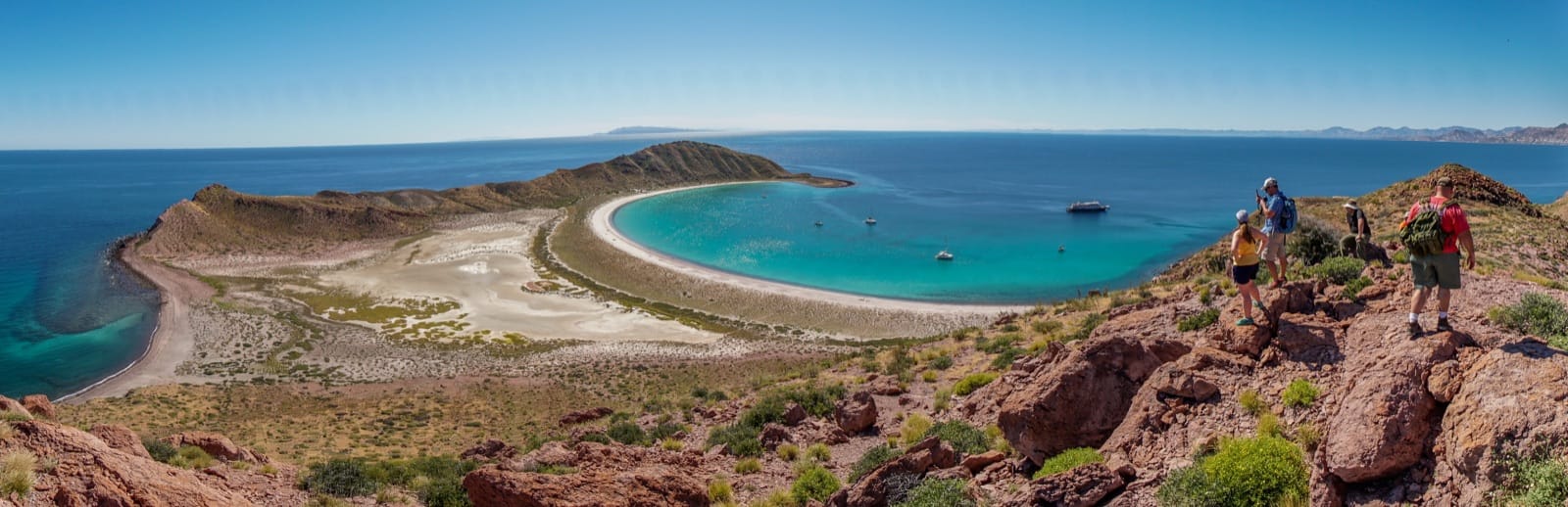 Panoramic view from the ridge above Isla San Francisco — two hikers standing on the rocky crest, looking down on the horseshoe-shaped beach and two small ships anchored in turquoise water.