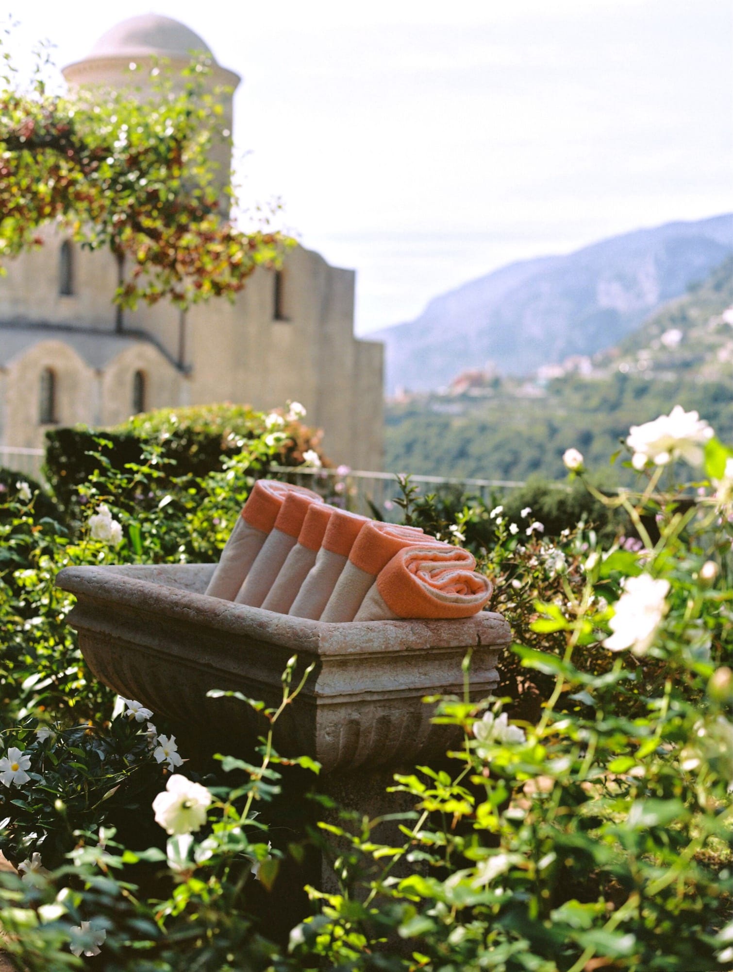 A garden seat at the Caruso — a single orange towel rolled on a stone bench, the Belvedere dome behind, the coast falling away past the roses.