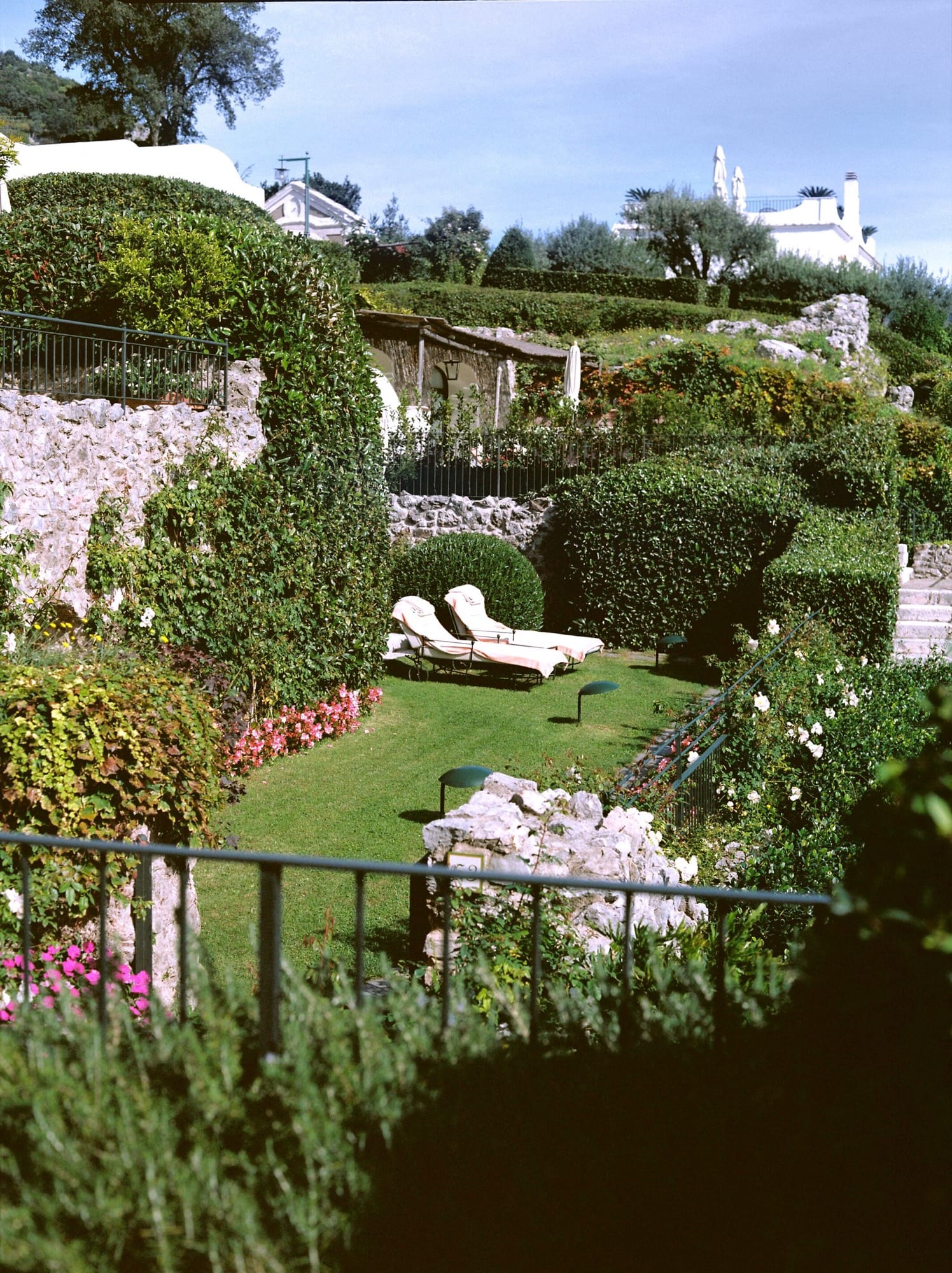 The Caruso garden terrace seen from above — clipped hedges, white loungers, stone, the village rooflines and coast beyond.