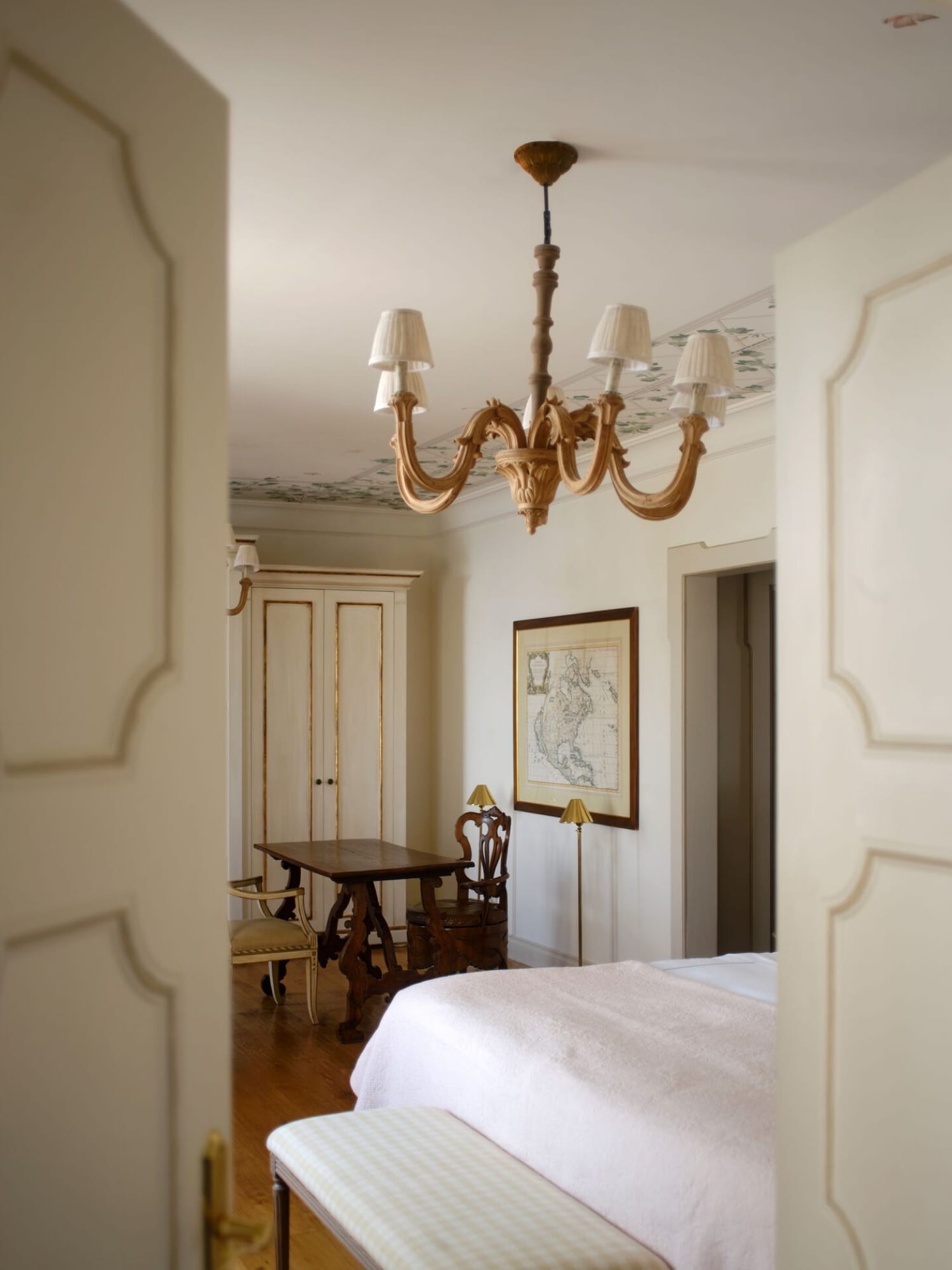 A Caruso bedroom seen through ornate painted double doors — a brass chandelier overhead, a glimpse of the bed beyond, an antique map on the wall.