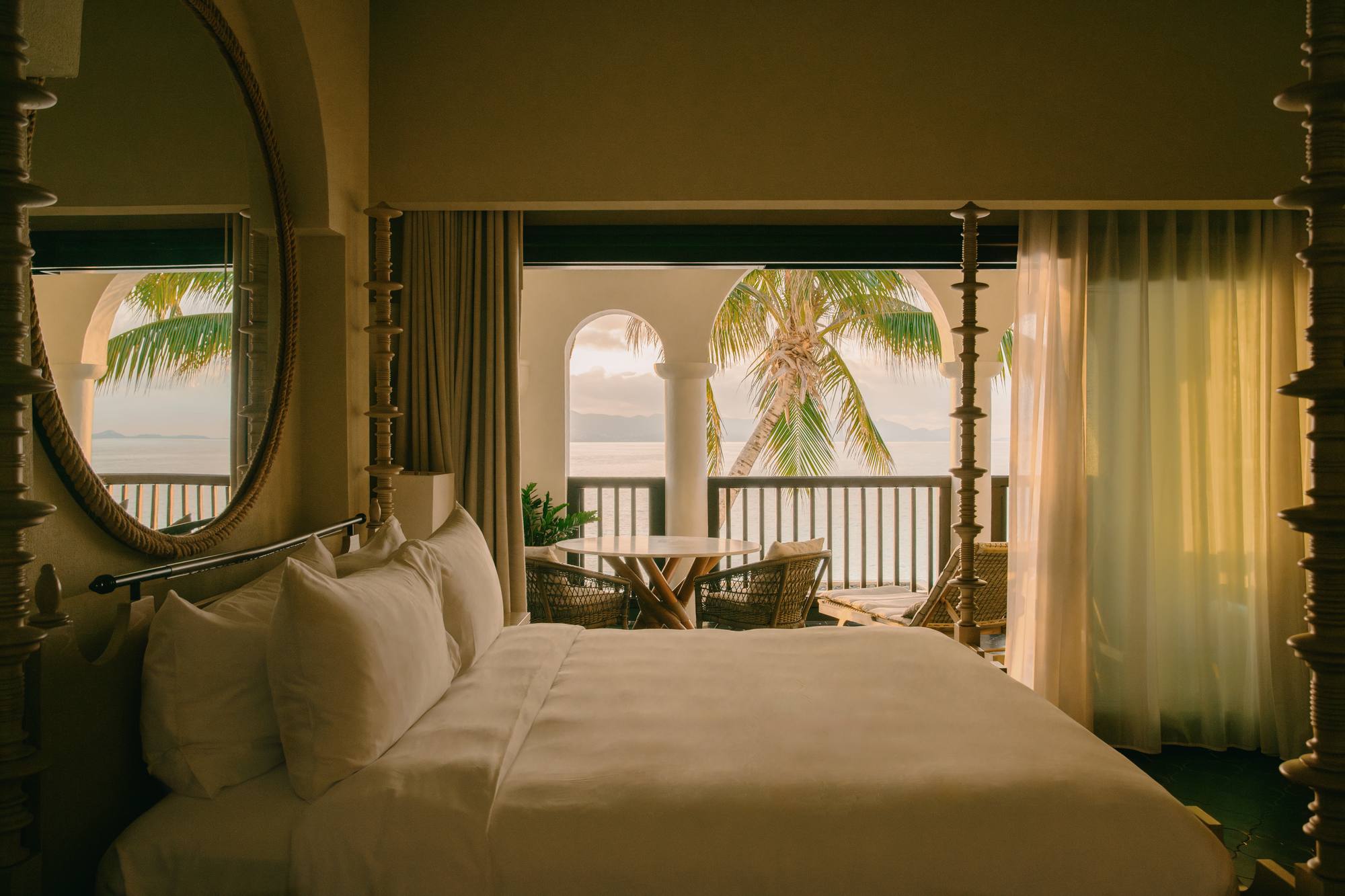 A cottage bedroom at Cap Juluca with a canopy bed, a round mirror, and an arched door opening onto a sea-view terrace.