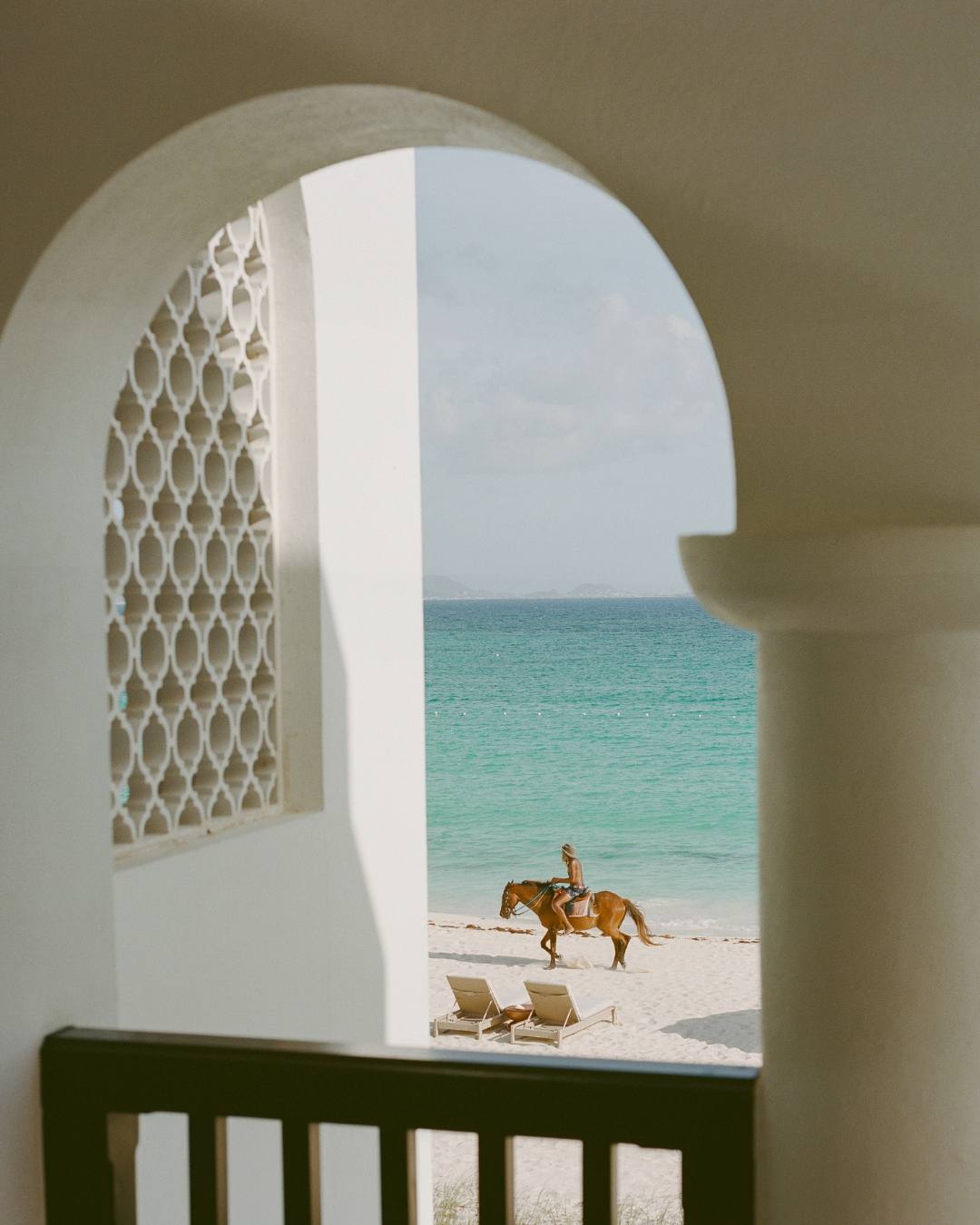 The view through a Greco-Moorish arched window at Cap Juluca, framing a rider on horseback along the empty beach.