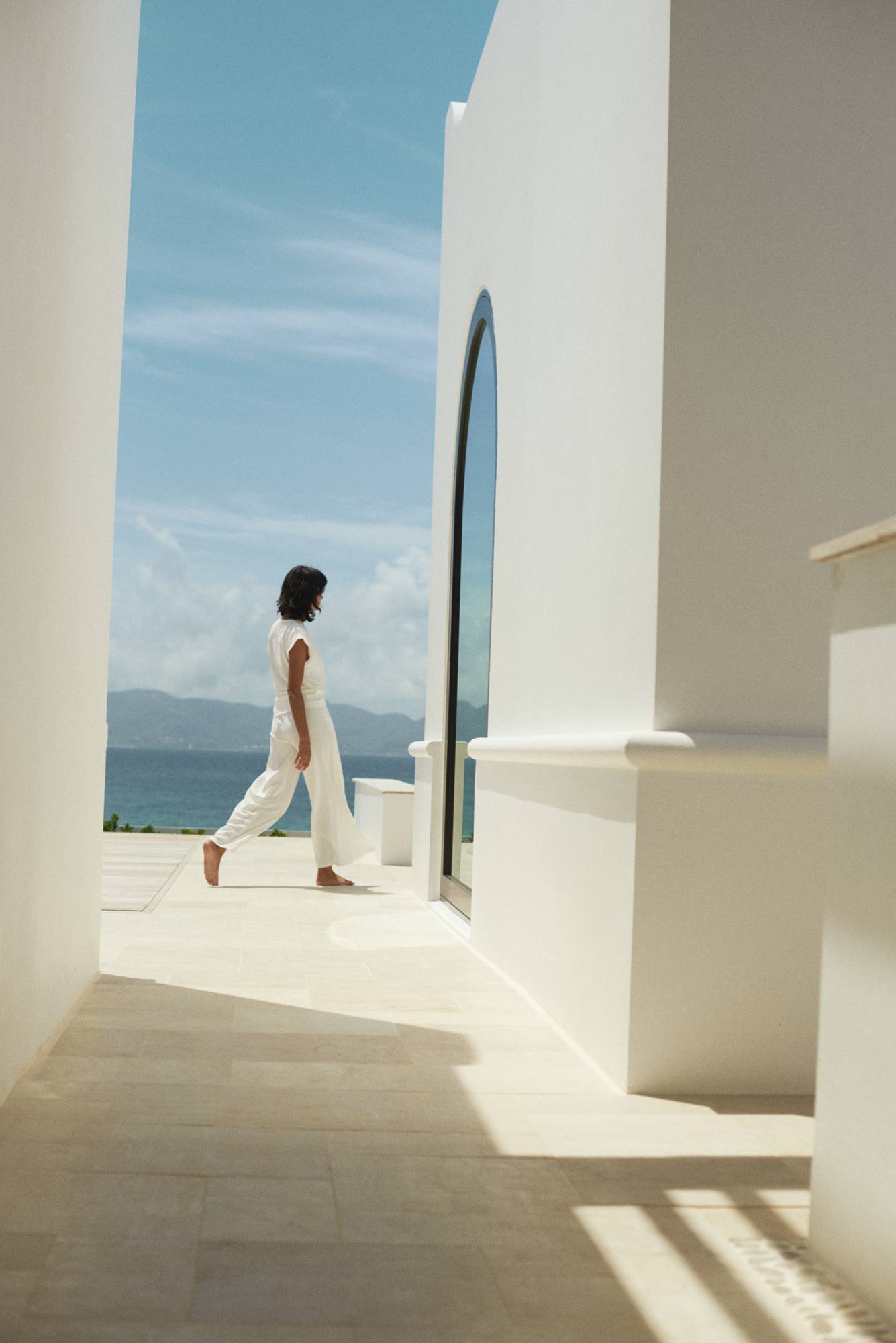 A figure in white linen walking a sunlit white-stone passage at Cap Juluca, the sea beyond.