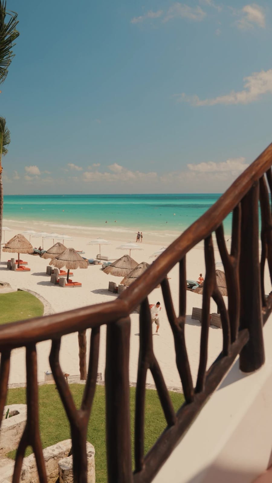 The beach at Maroma seen through a wooden casita pergola railing — thatched palapa umbrellas in the white sand, the turquoise Caribbean horizon, a guest crossing the sand toward the water.