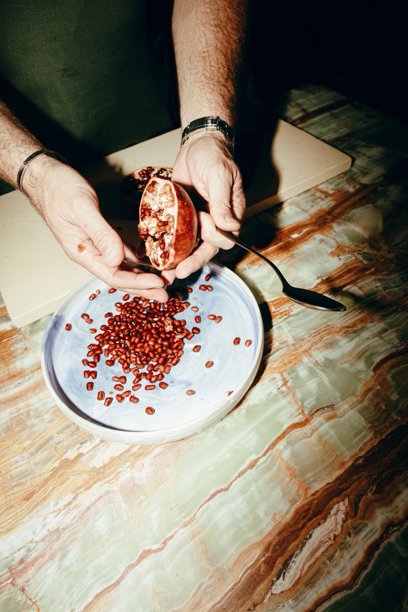 Hands cracking a pomegranate over a blue-and-white ceramic plate on an onyx-marble table at Maroma, ruby seeds spilling onto the dish.