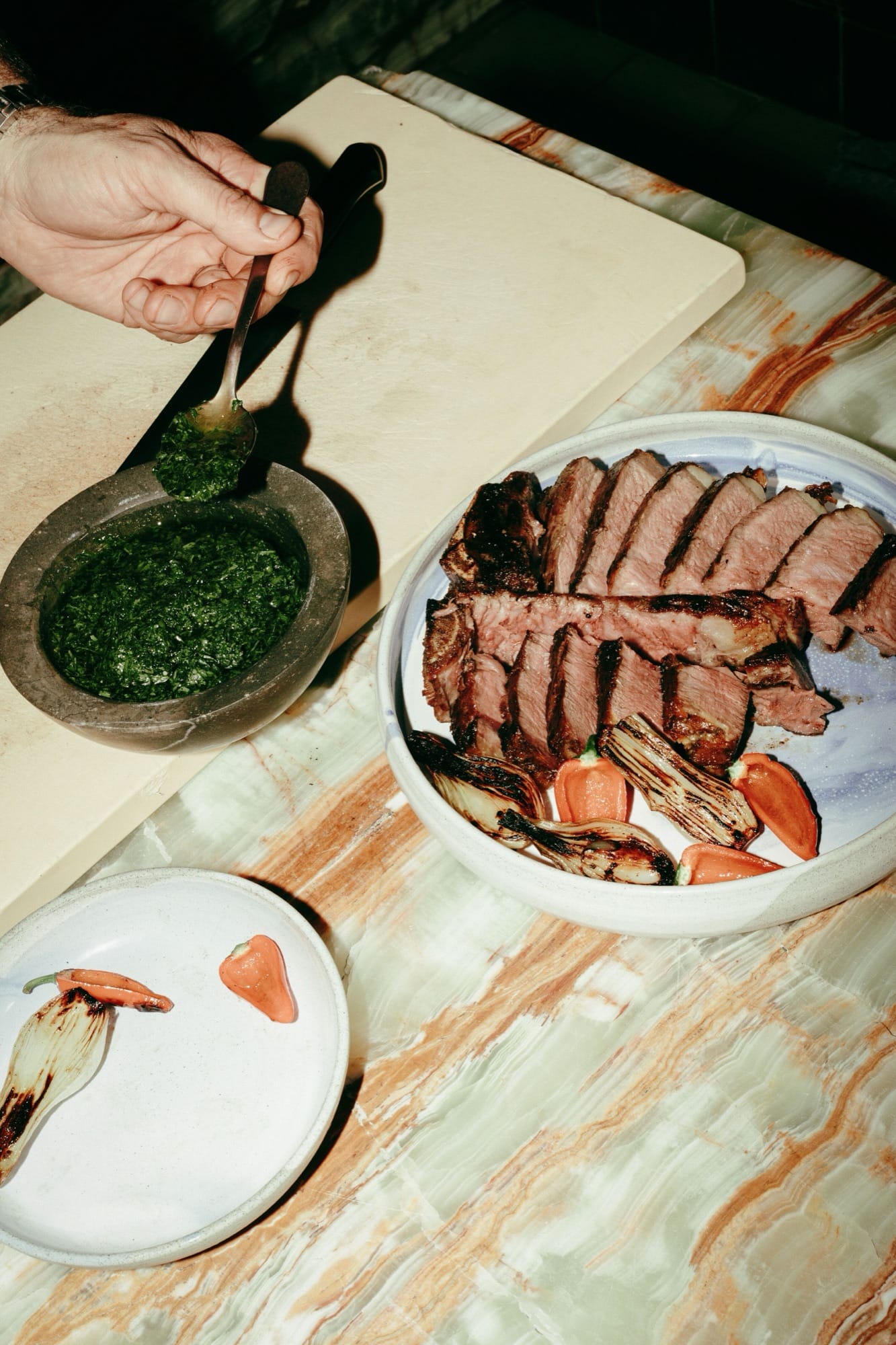 A hand spooning fresh chimichurri over a sliced ribeye on a turquoise-glazed Mexican ceramic platter — charred scallions and tomatoes alongside, the green stone-mortar at the edge of the marble table.