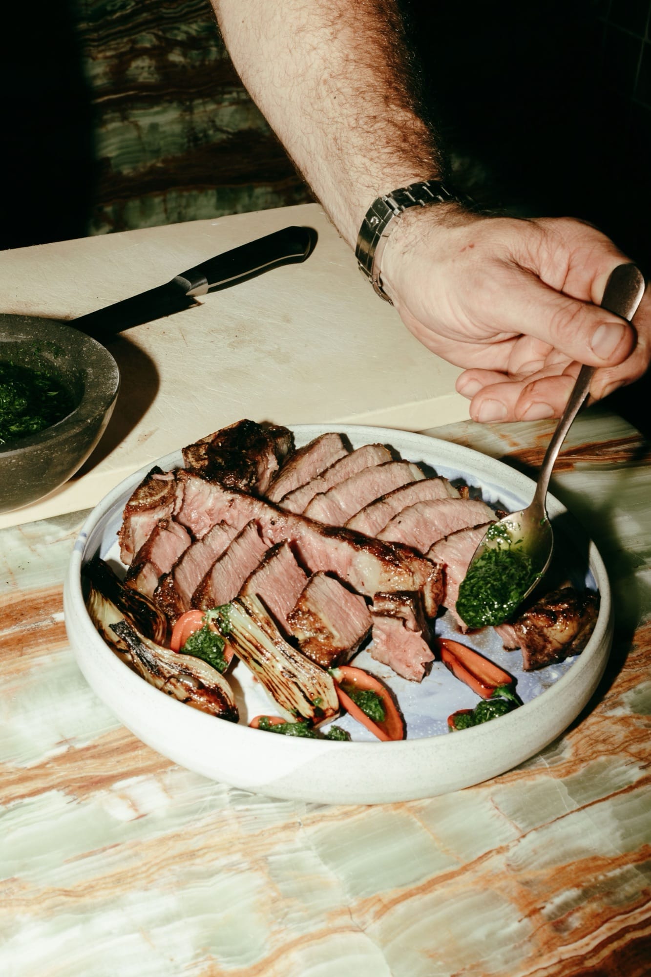 A close-in plating shot of the chimichurri-and-ribeye dish at Maroma — the green sauce spooned onto pink-medium slices, charred scallion and tomato around the rim.