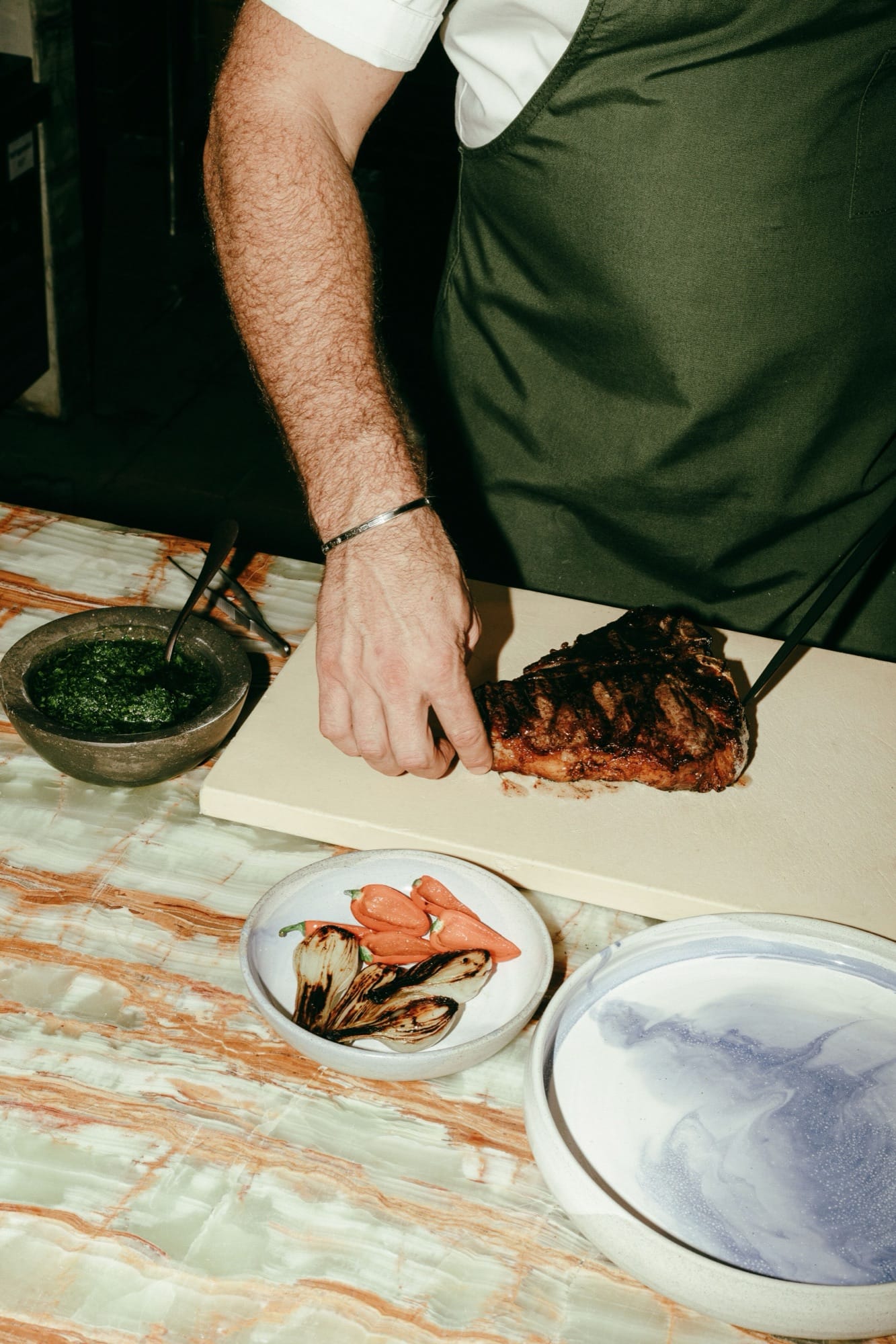 A Maroma chef in white coat and dark green apron setting a charred ribeye onto a marble cutting board, a small bowl of fresh chimichurri and a dish of roasted tomatoes on the side.