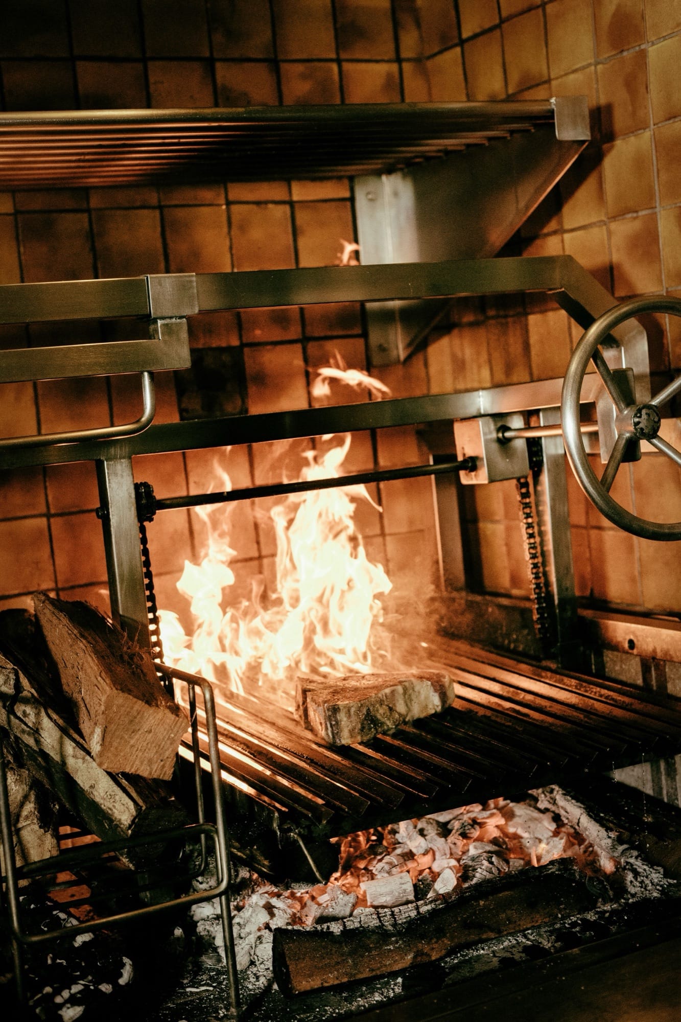 The open-flame grill at Maroma — a tile-back hearth with iron rails, flame licking up over the grate, charred wood and embers below.