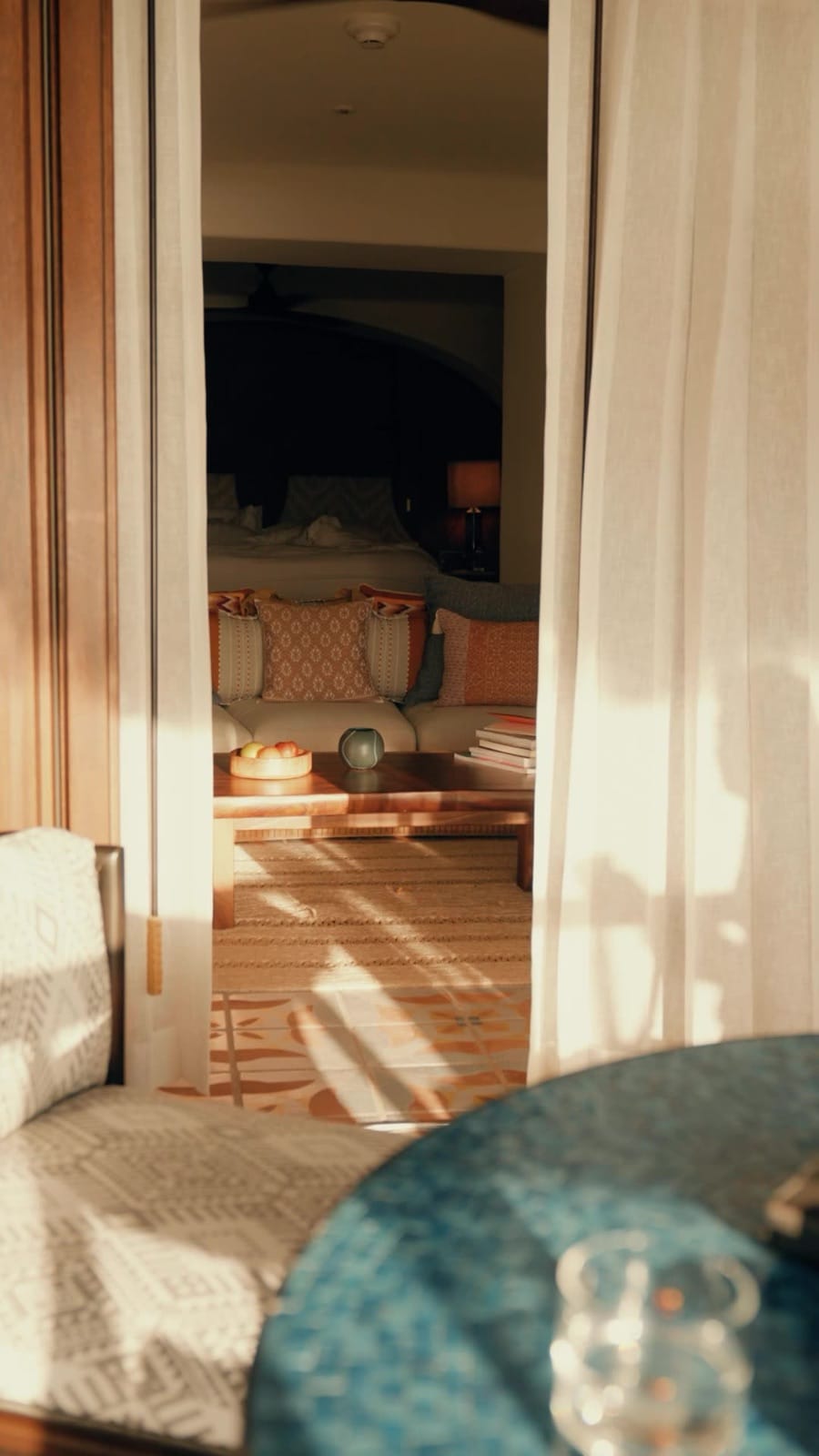 Through-the-curtain view into a Maroma casita sitting room — terra-cotta tiled floor with patterned inlay, daybed with kilim throw and patterned pillows, low coffee table with a bowl of fruit, the bedroom and arched doorway in soft shadow.