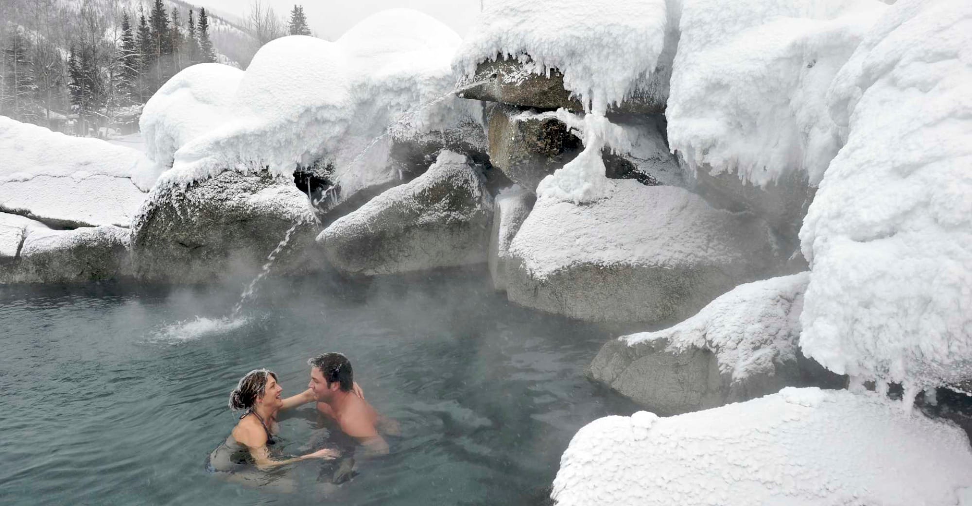 A couple soaking in the steaming outdoor rock pool at Chena Hot Springs, surrounded by snow-laden boulders.