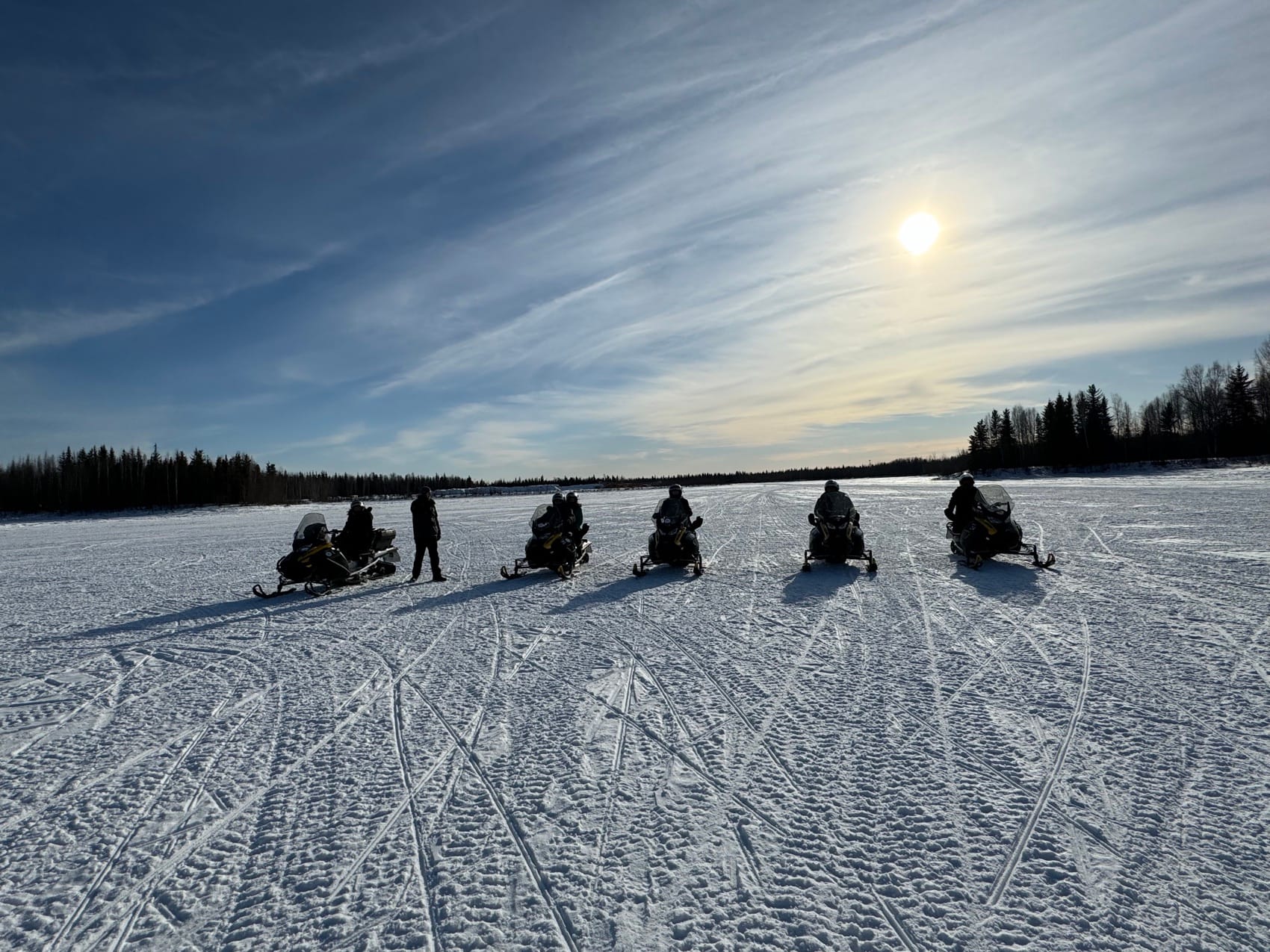 A bundled-up traveler grinning on a snowmobile on a groomed forest trail under bright blue sky.
