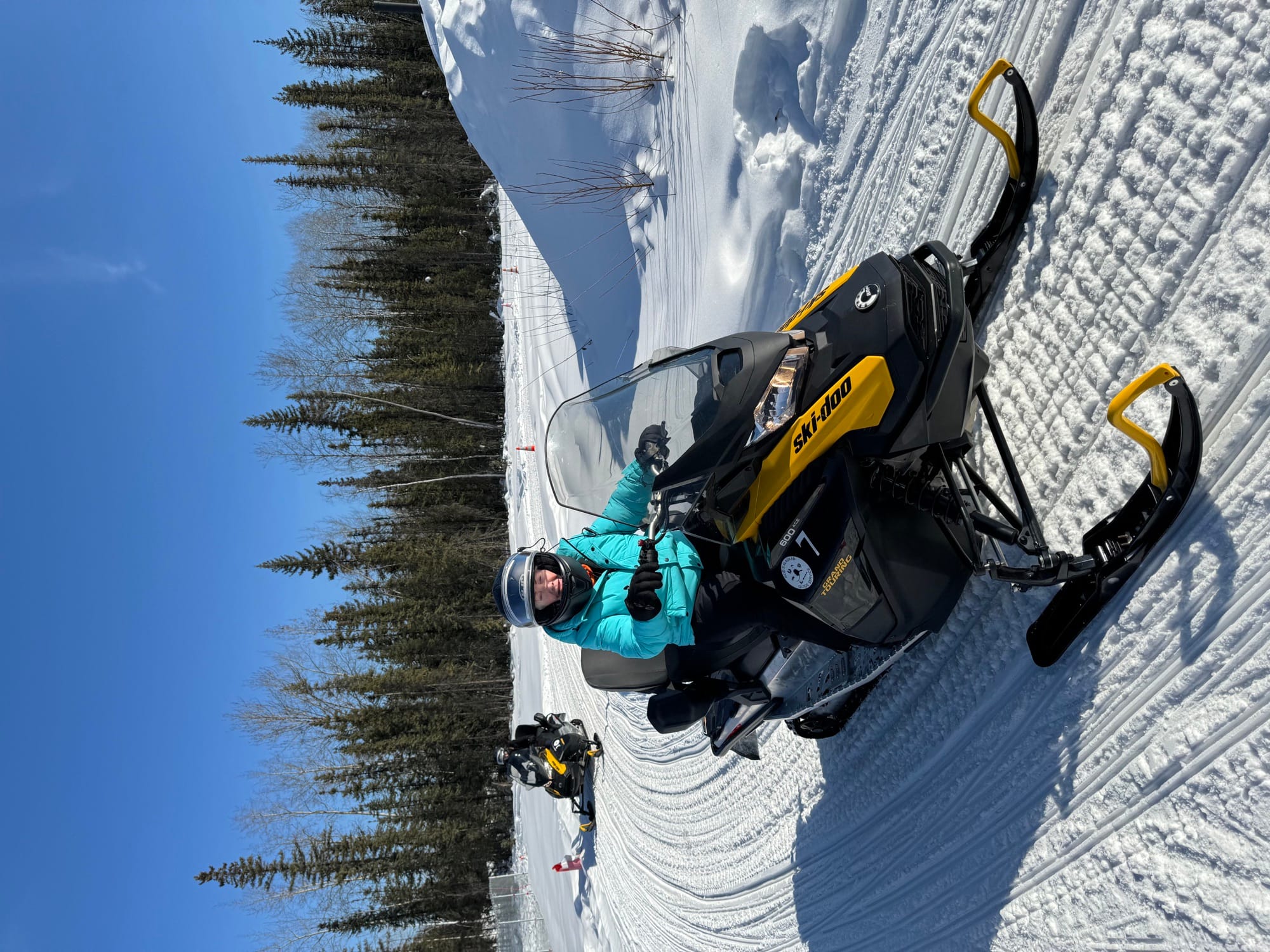 A line of guided snowmobiles paused on a frozen, sunlit lake ringed by boreal forest.