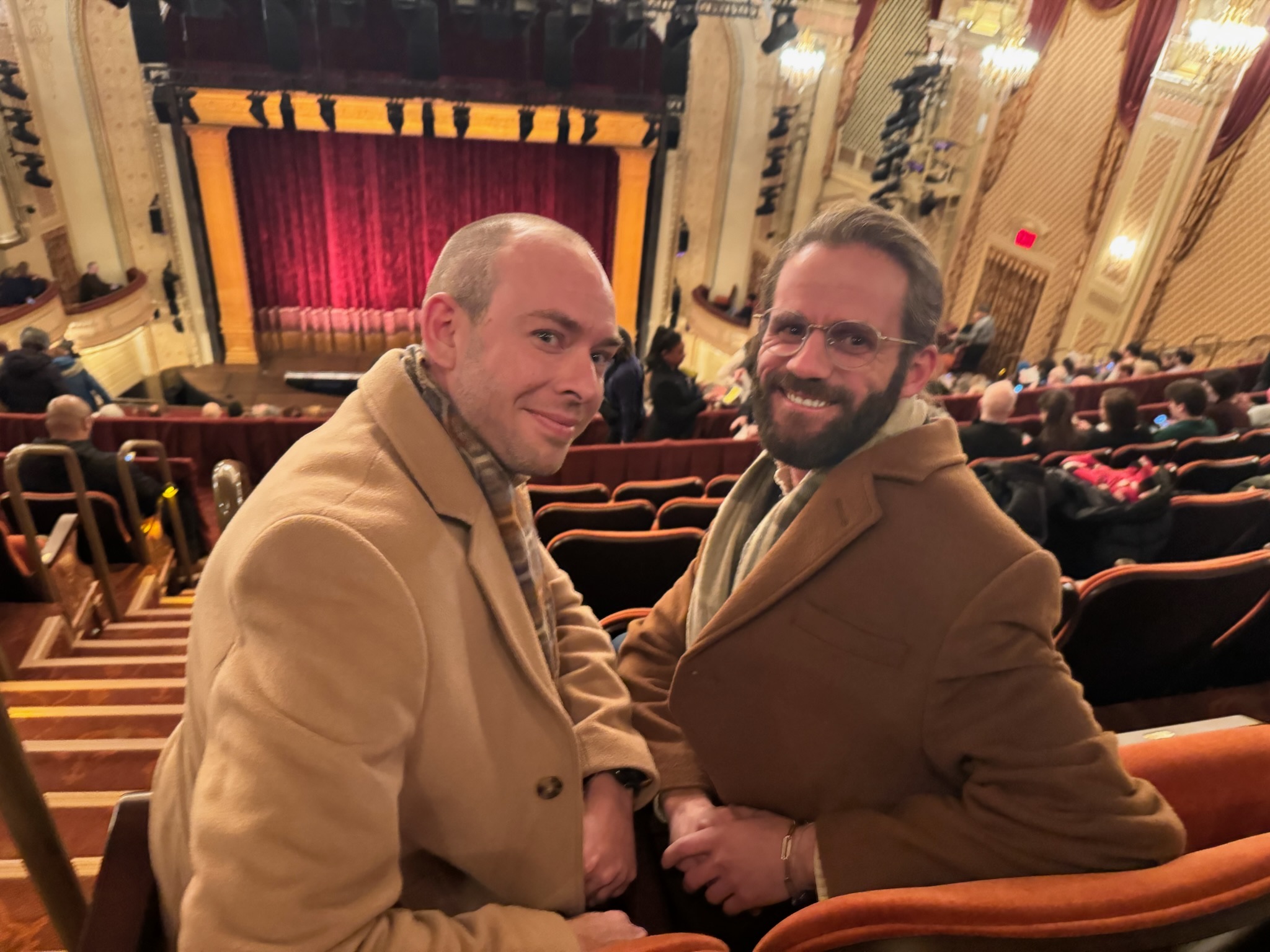 Erik and his husband Chase in their overcoats, seated in a gilded Broadway theatre before the curtain goes up.