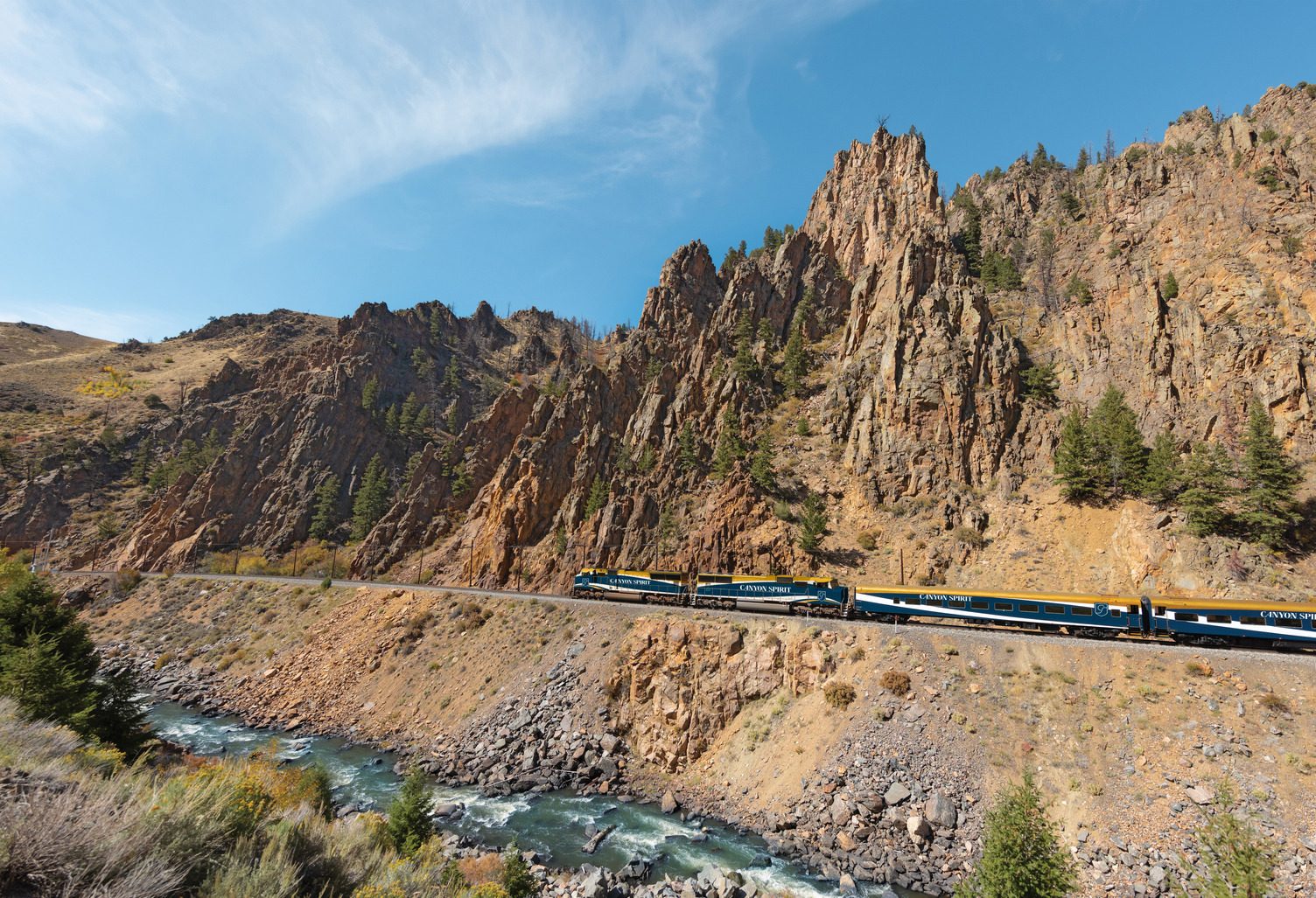The Canyon Spirit train rounds a bend below golden rock spires, a river running beside the track.