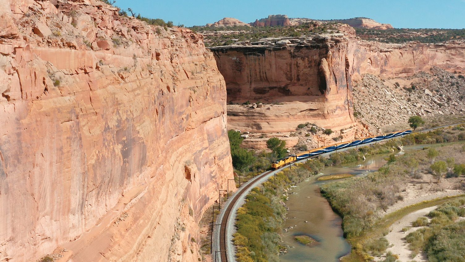 A glass-dome train threads a deep red sandstone canyon beside a river on the Rockies to the Red Rocks route.