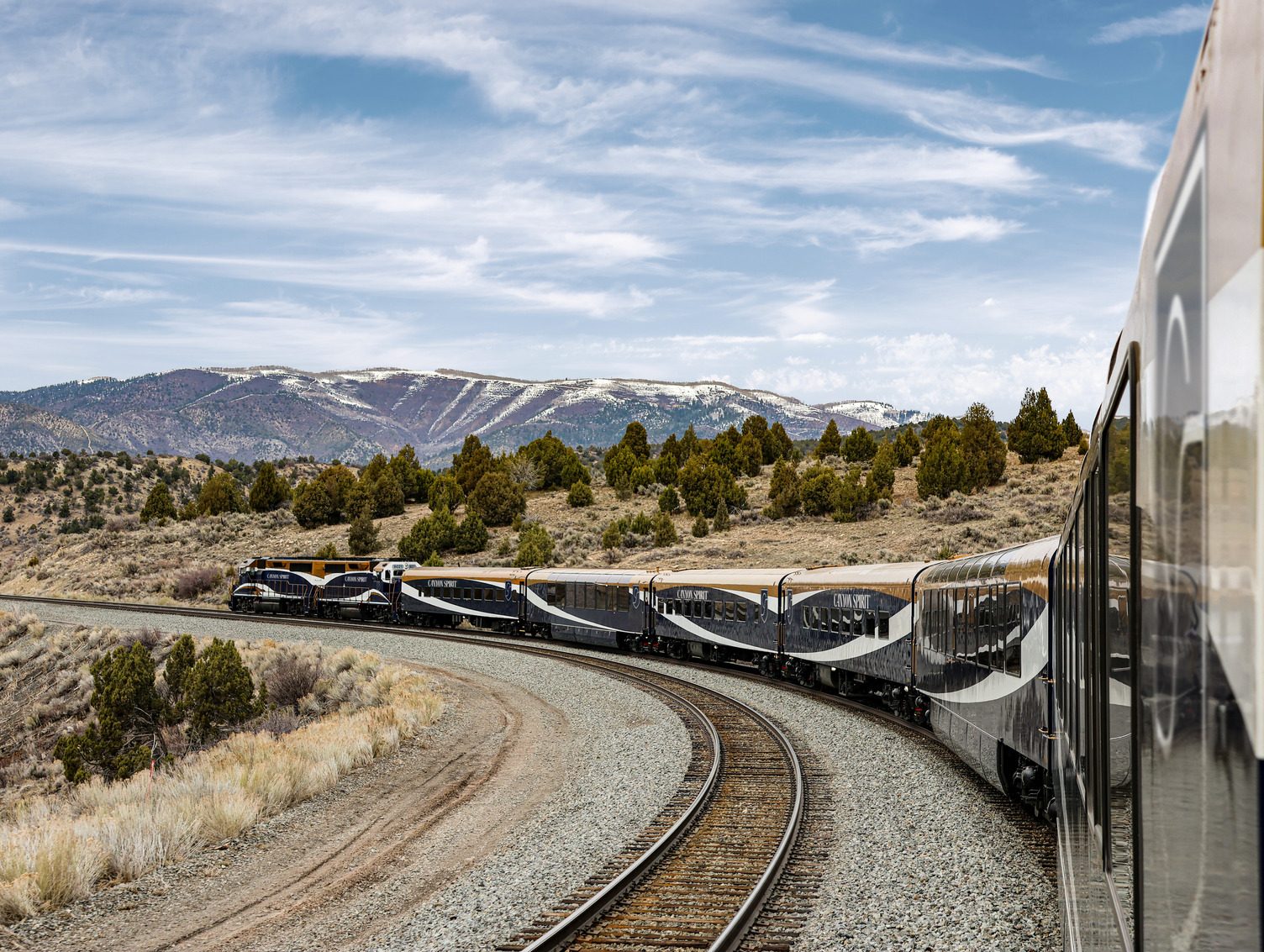 The Canyon Spirit train curves through high-desert sage country with snow-dusted peaks on the horizon.