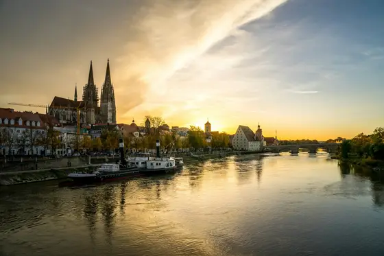 The Danube at Regensburg, Germany, at golden hour — cathedral spires, the medieval Stone Bridge, and a moored river vessel.