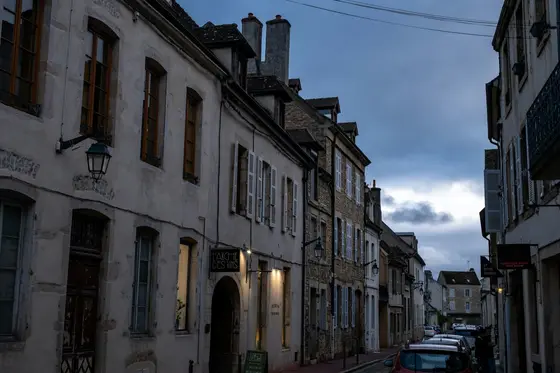 A quiet cobblestone street at dusk in Beaune, Burgundy — warm light glowing in the windows of old stone buildings.