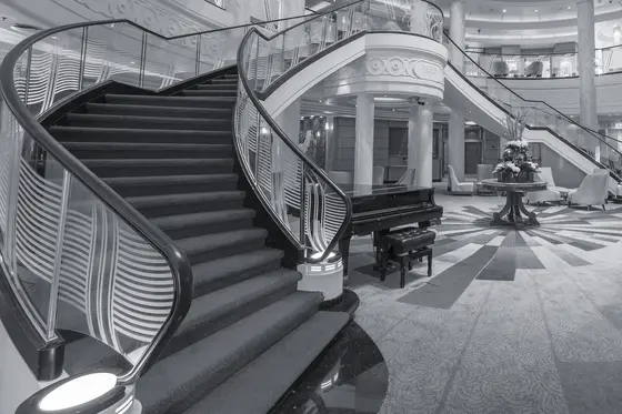 The Grand Lobby of Queen Mary 2 in black and white: a sweeping staircase with etched-glass balustrades, a grand piano at its foot, the lobby floor radiating out beyond.