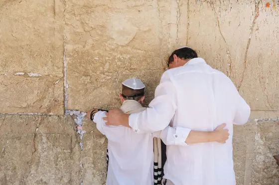 A father in white linen holds his son close at the Western Wall in Jerusalem; both wear kippot, the son in tefillin, prayer notes tucked into the limestone seams.