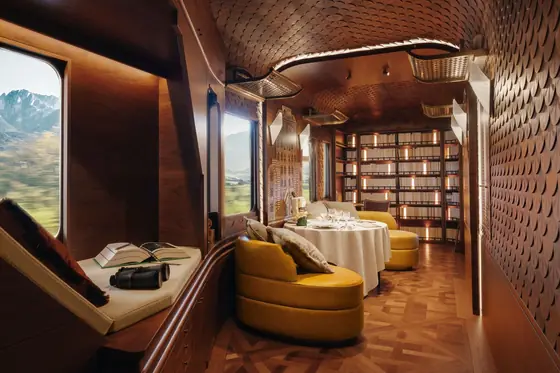 The interior of L'Observatoire on the Venice Simplon-Orient-Express — marquetry panels, a yellow banquette, a table laid for dinner, and a book left open on the window seat.
