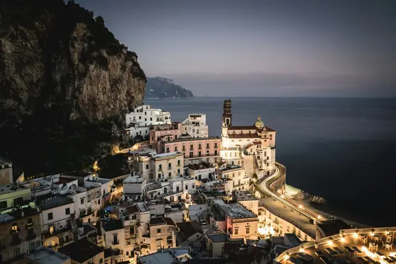 Atrani at dusk, lights coming on in stacked pastel houses folded into the cliff above a small beach.