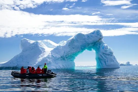 A zodiac of passengers in red parkas passing beneath a blue arch of iceberg on the Antarctic Peninsula.