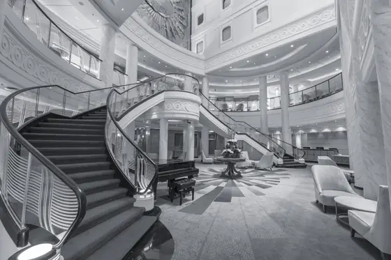 The Grand Lobby of Queen Mary 2 in black and white — a double staircase curving down to a marble floor, a grand piano at its foot, under a domed ceiling.