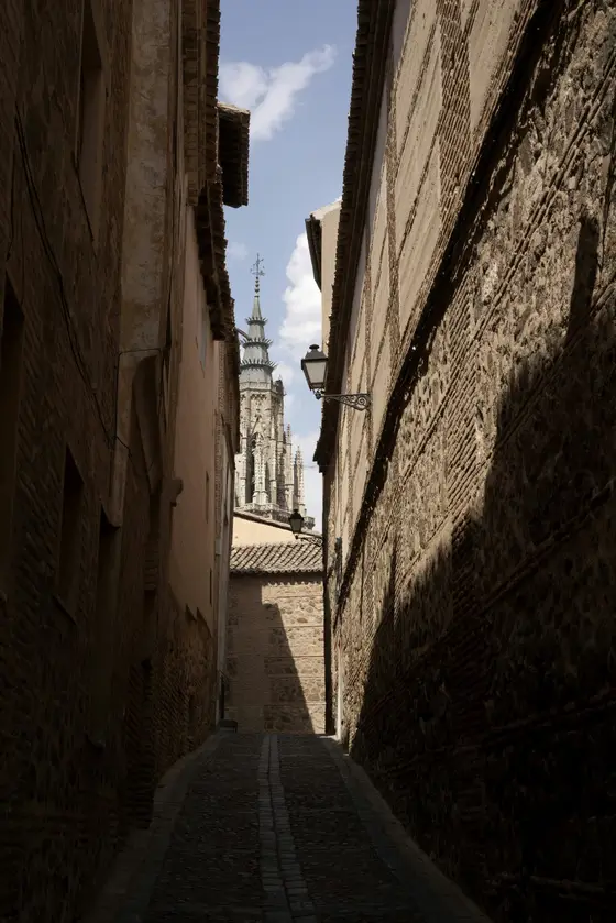 A narrow, empty stone lane climbs through Toledo's old town toward a distant bell tower — the kind of quiet alley that threads the city's medieval Judería.