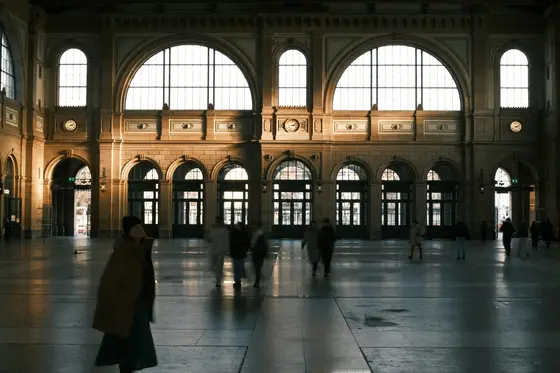 A grand, sunlit train station hall with travelers passing through — the great terminus where timing is everything.