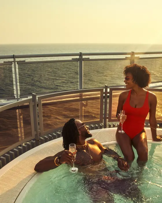 A couple sharing champagne in the hot tub at Richard's Rooftop aboard a Virgin Voyages ship, the ocean glowing behind them at sunset.