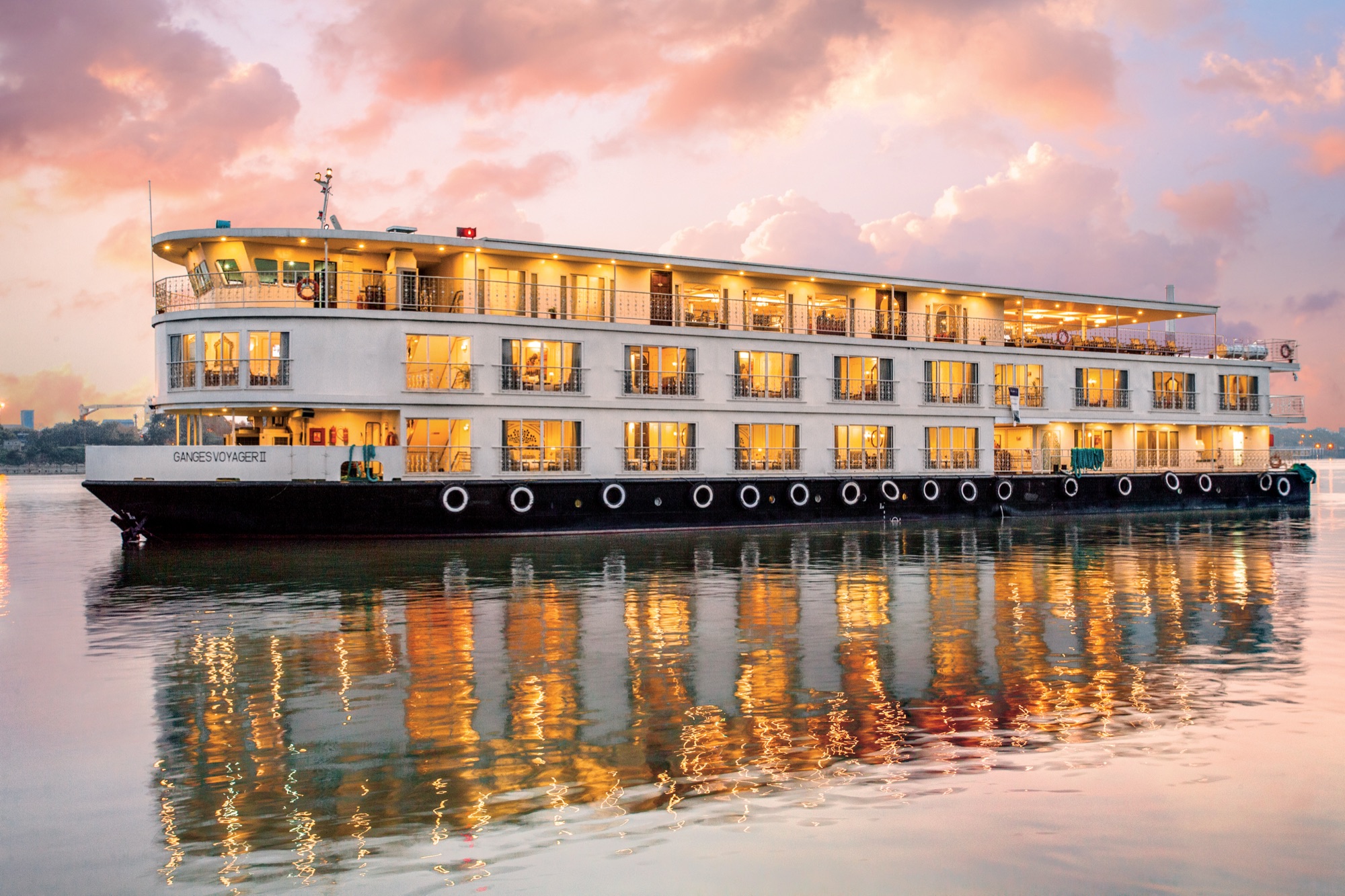 The Ganges Voyager II lit at dusk on the Ganges, its windows reflecting gold in the water under a pink sky.