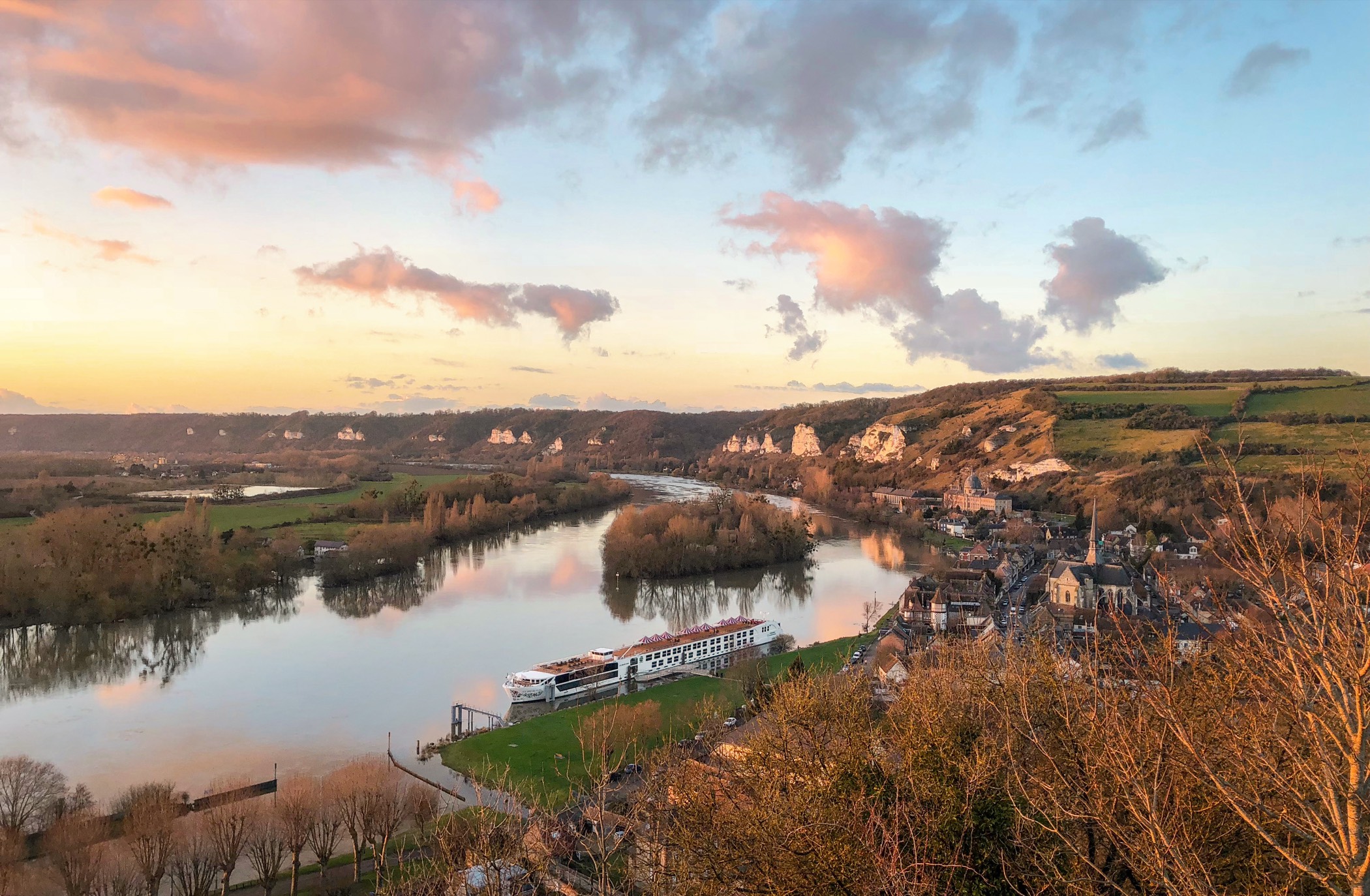 A Uniworld Super Ship moored on the Seine below a French village at sunset — the S.S. Joie de Vivre in the golden light of the river valley.